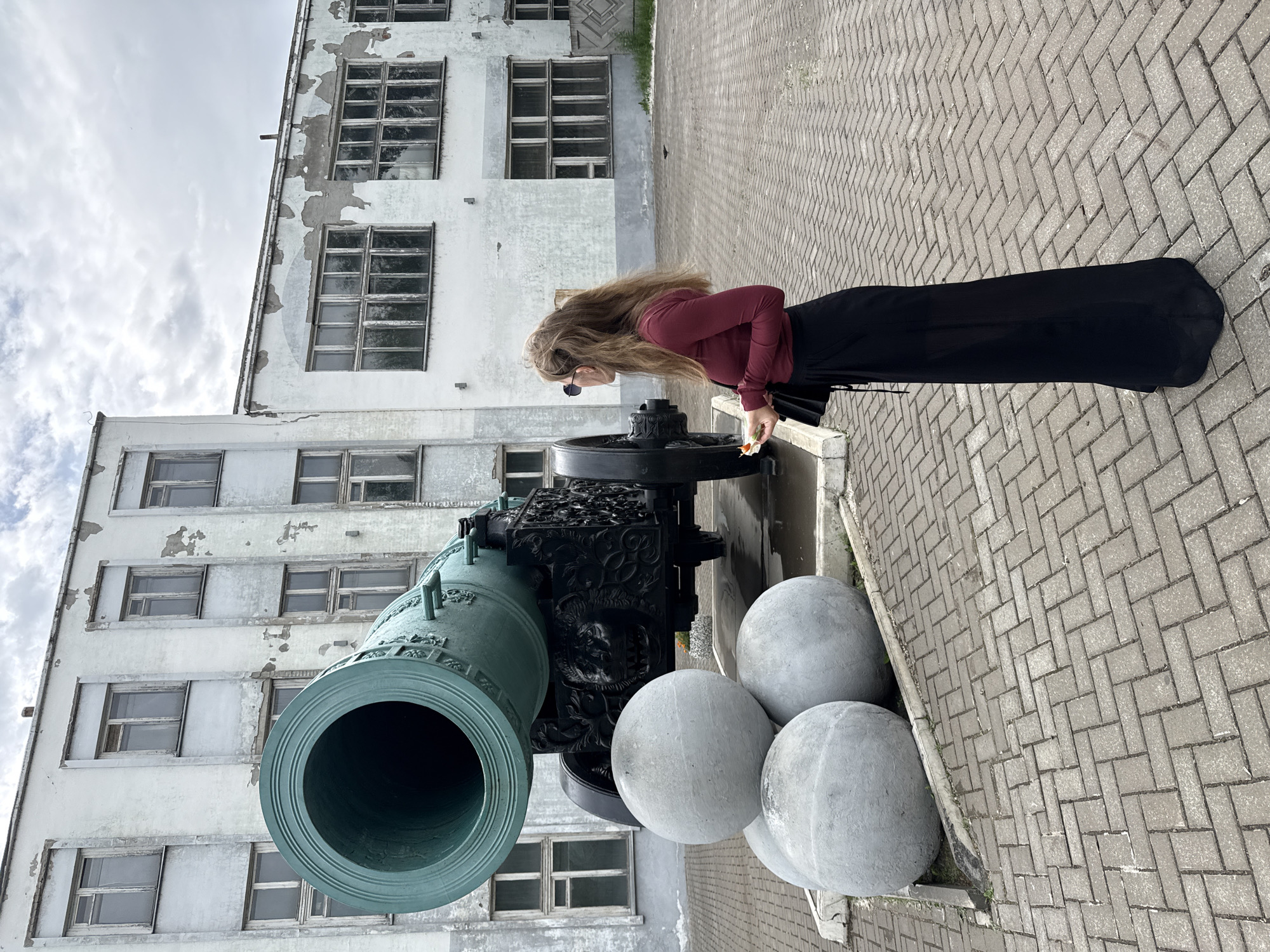 In Izhevsk, Russia, an outdoor scene features a vintage cannon with three large cannonballs in front of a weathered, white building. A young woman with long, light brown hair, wearing a maroon long-sleeved shirt and a long black skirt, stands to the right of the cannon. She is looking towards the cannon, holding a small snack in her right hand and a black purse in her left. The cannon is mounted on a dark, ornate carriage with decorative elements, including a lion's head carving. The building behind them has multiple windows and shows signs of peeling paint. The ground is paved with grey interlocking bricks. The sky is cloudy.