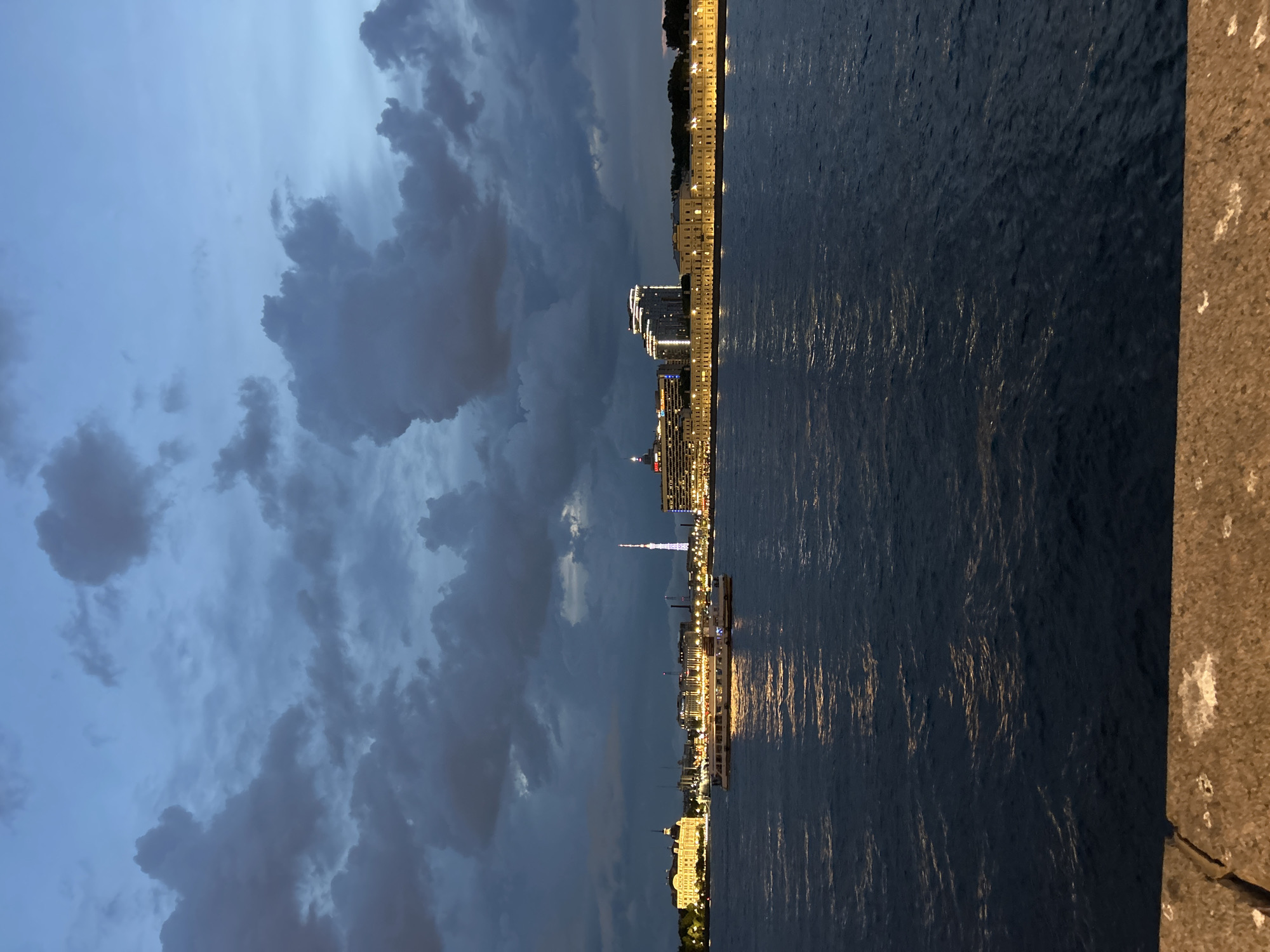 An outdoor, wide-angle, slightly elevated perspective captures a city riverfront scene at dusk or night, dominated by a dark, overcast sky and a wide body of water. The upper portion of the image is filled with a stormy, dark blue-grey sky featuring numerous large, amorphous clouds.
The midground is occupied by a dark blue river with moderate ripples, reflecting artificial lights as shimmering streaks of yellow and orange. Two white passenger boats, illuminated from within, are visible on the water, generating prominent reflections. The boat on the left is slightly larger and positioned closer to the viewer.
The far bank features a distinct city skyline. From left to right: a large, multi-story, light-colored building with a domed roof and numerous illuminated windows; a cluster of modern buildings, some with lit facades; a tall, slender television tower glowing with purple and blue lights, serving as a central vertical element; further to the right, a modern high-rise building with a stepped roofline and bright white illumination, bearing a visible "CITYTEL" sign; and finally, a long, continuous block of light-colored, classically styled buildings, uniformly illuminated along their facades, extending into the distance on the right. Lush green trees are visible behind some of the buildings on the right bank.
The immediate foreground reveals a rough-textured, brown-grey stone embankment or wall, partially lit, with visible cracks and light-colored patches (possibly remnants of bird droppings or worn concrete) along its surface. The vantage point appears to be from another embankment overlooking the river.
The location is the Neva River in St. Petersburg, Russia, with the Saint Petersburg Television Tower prominent in the background and characteristic city architecture lining the banks.