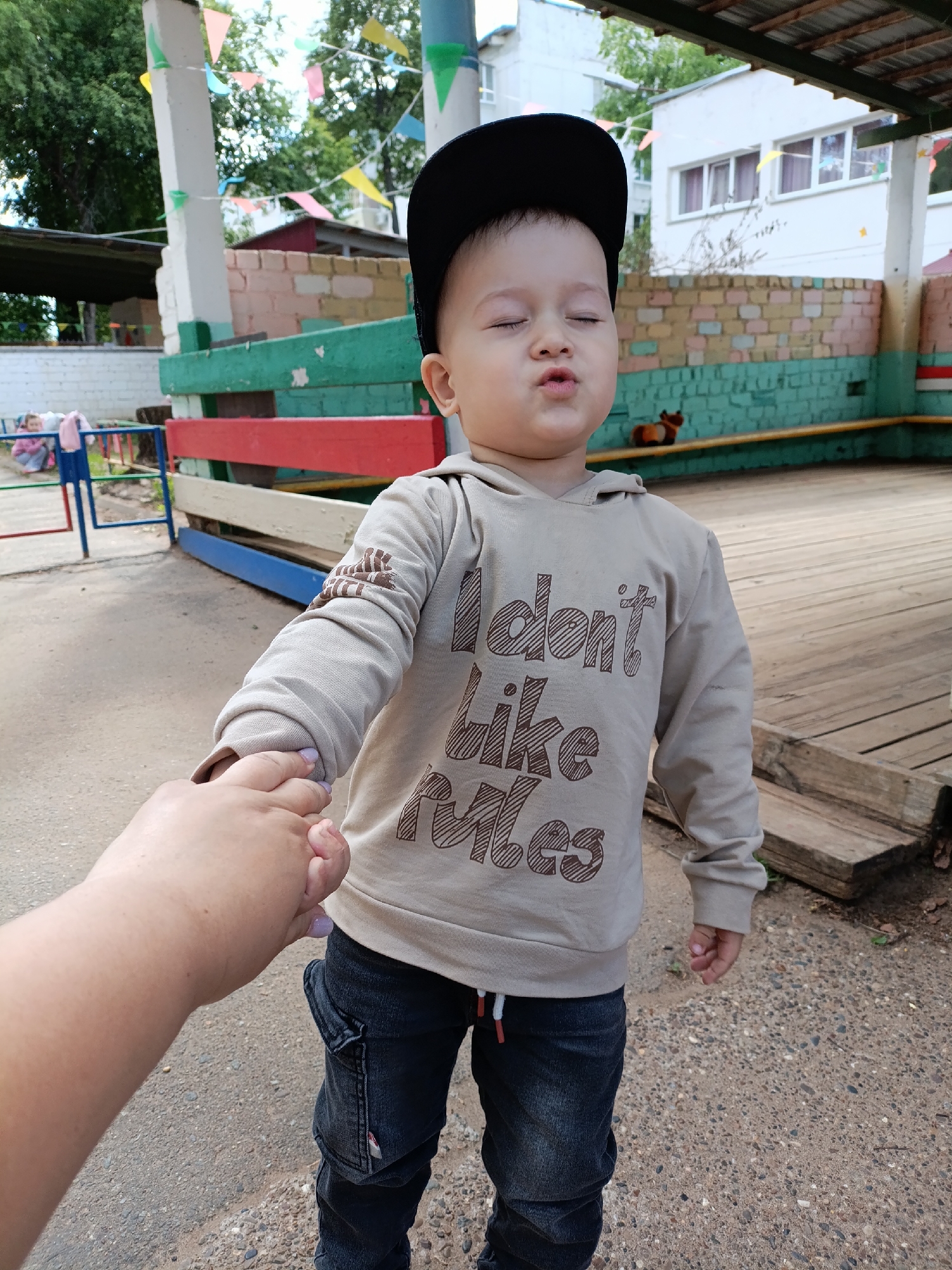 A young child wearing a gray hooded sweatshirt with the text "I don't like rules" and a black baseball cap is holding hands with an adult. The child's eyes are closed, and their lips are pursed. The adult's hand is visible, with fingernails painted a light purple. The setting appears to be an outdoor play area with colorful fences, a wooden deck, and a building in the background. Based on information available, the location is identified as the city of Chaikovsky, Russia.