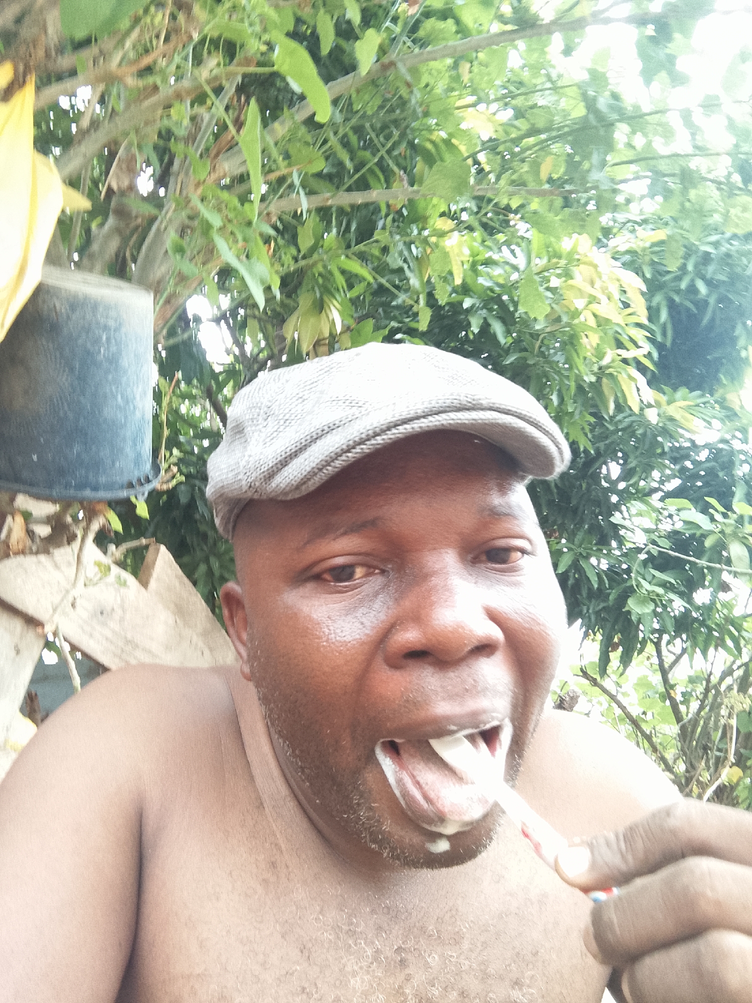A shirtless man wearing a gray cap is brushing his teeth outdoors. He is holding a toothbrush with white toothpaste in his mouth. Behind him, there is lush green foliage and a black bucket hanging from a wooden structure. The location is Unknown, Unknown.