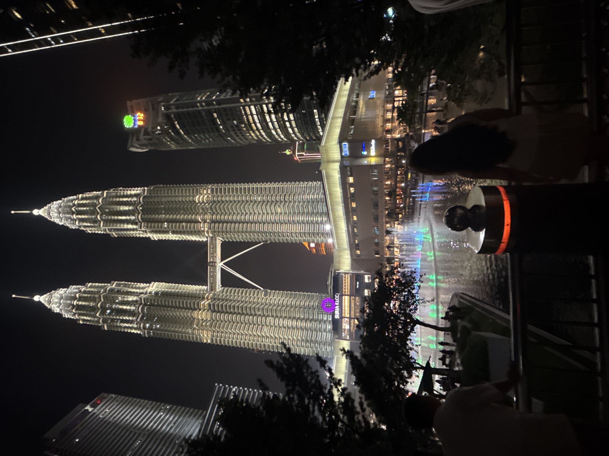 The image captures a night view of Kuala Lumpur, Malaysia, showcasing the Petronas Twin Towers and a nearby skyscraper. The towers are illuminated, with their distinctive stacked design clearly visible. A connecting skybridge links the two towers. In front of the towers, there appears to be a fountain, illuminated with green and blue lights. Several people can be seen in the foreground, silhouetted against the lit cityscape. There's a telescope-like object in the foreground. The overall environment suggests a tourist spot or public viewing area.