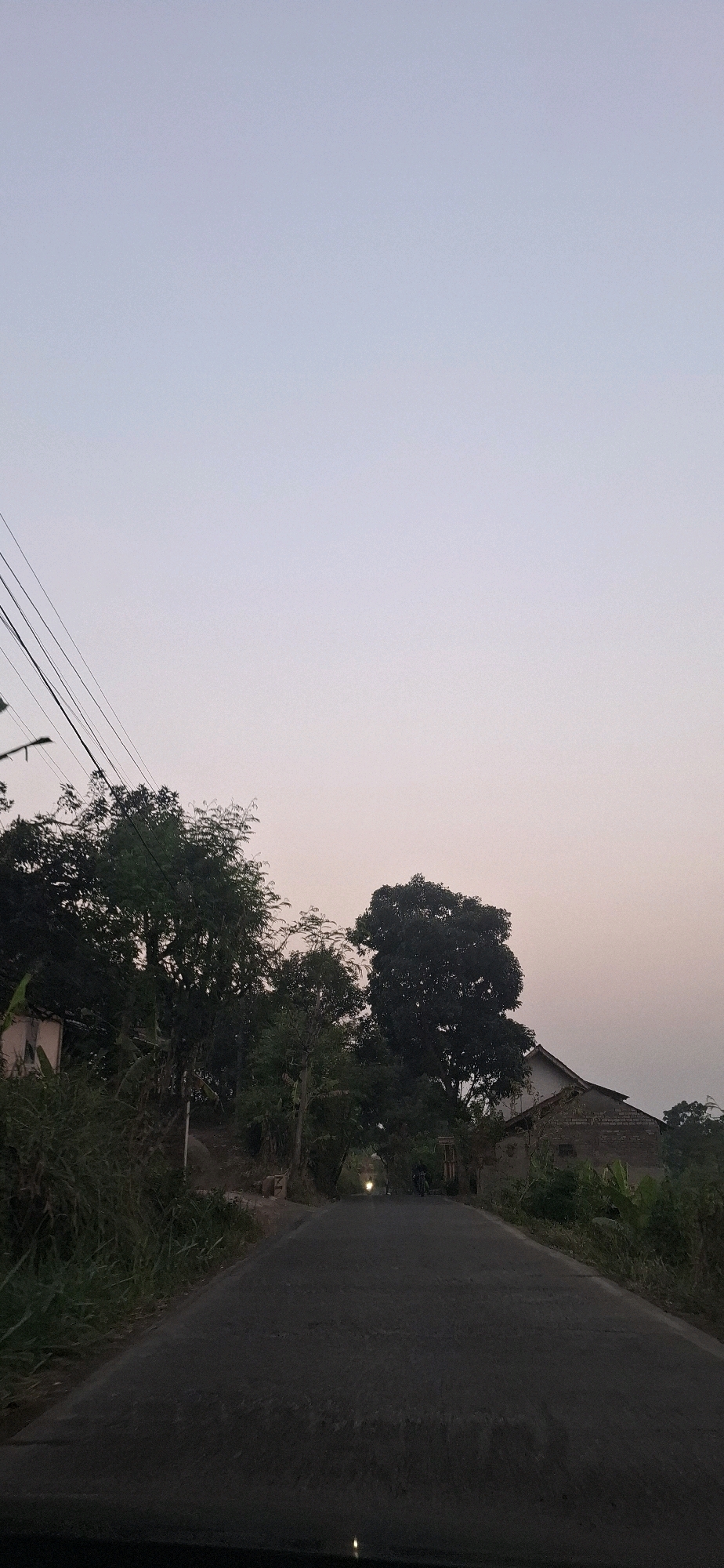 The image depicts a paved road in a rural or suburban setting, possibly Cibinong Hilir, Indonesia, during twilight or early morning hours. The sky is a pale, gradient of light blue and pink, suggesting low light conditions. On the left side of the road, there are dense trees and vegetation, with a light-colored structure partially visible behind the foliage. Power lines are strung across the upper left portion of the image. On the right side, a simple brick house with a pitched roof is situated amongst lush greenery, including what appears to be banana plants. The road itself is dark gray asphalt, with some texture visible. In the distance, a single bright light, likely from a motorcycle, is visible on the road, indicating movement or a stationary vehicle. The overall atmosphere is quiet and serene, characteristic of a rural landscape at dusk or dawn.