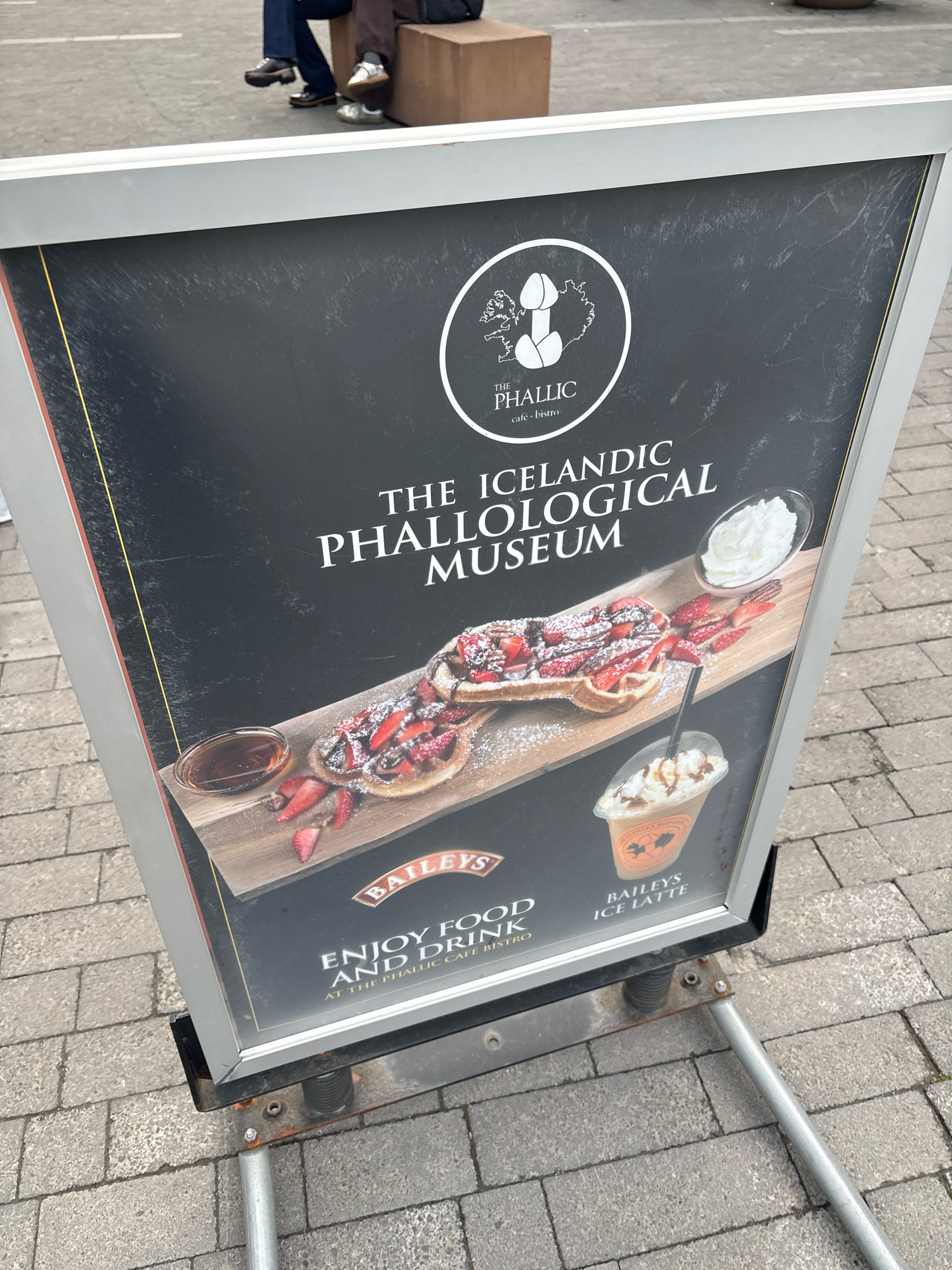 An A-frame sign stands on a sidewalk composed of grey pavers in Reykjavik, Iceland. The sign advertises "The Icelandic Phallological Museum" and "The Phallic Cafe Bistro". The sign features a logo with a stylized phallus and the outline of Iceland. Below the logo, the text reads "THE ICELANDIC PHALLOLOGICAL MUSEUM". A photograph of waffles with strawberries and whipped cream, a cup of chocolate sauce, and a Baileys Ice Latte is displayed on the lower half of the sign. Text at the bottom of the sign reads "ENJOY FOOD AND DRINK AT THE PHALLIC CAFE BISTRO" and "BAILEYS ICE LATTE". In the background, a person in dark pants and shoes sits on a low wooden bench, with another person's legs visible to their left.