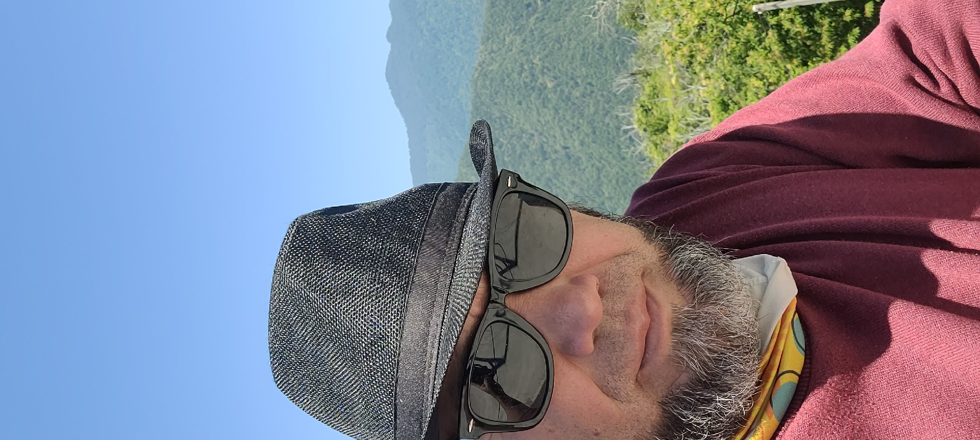 A medium shot is captured of a person wearing a black fedora and black sunglasses, facing the camera. The subject has graying facial hair and is wearing a maroon shirt with a colorful bandana around their neck. The background features a lush, green, mountainous landscape under a clear blue sky. This scene is located in Milisi, Georgia.