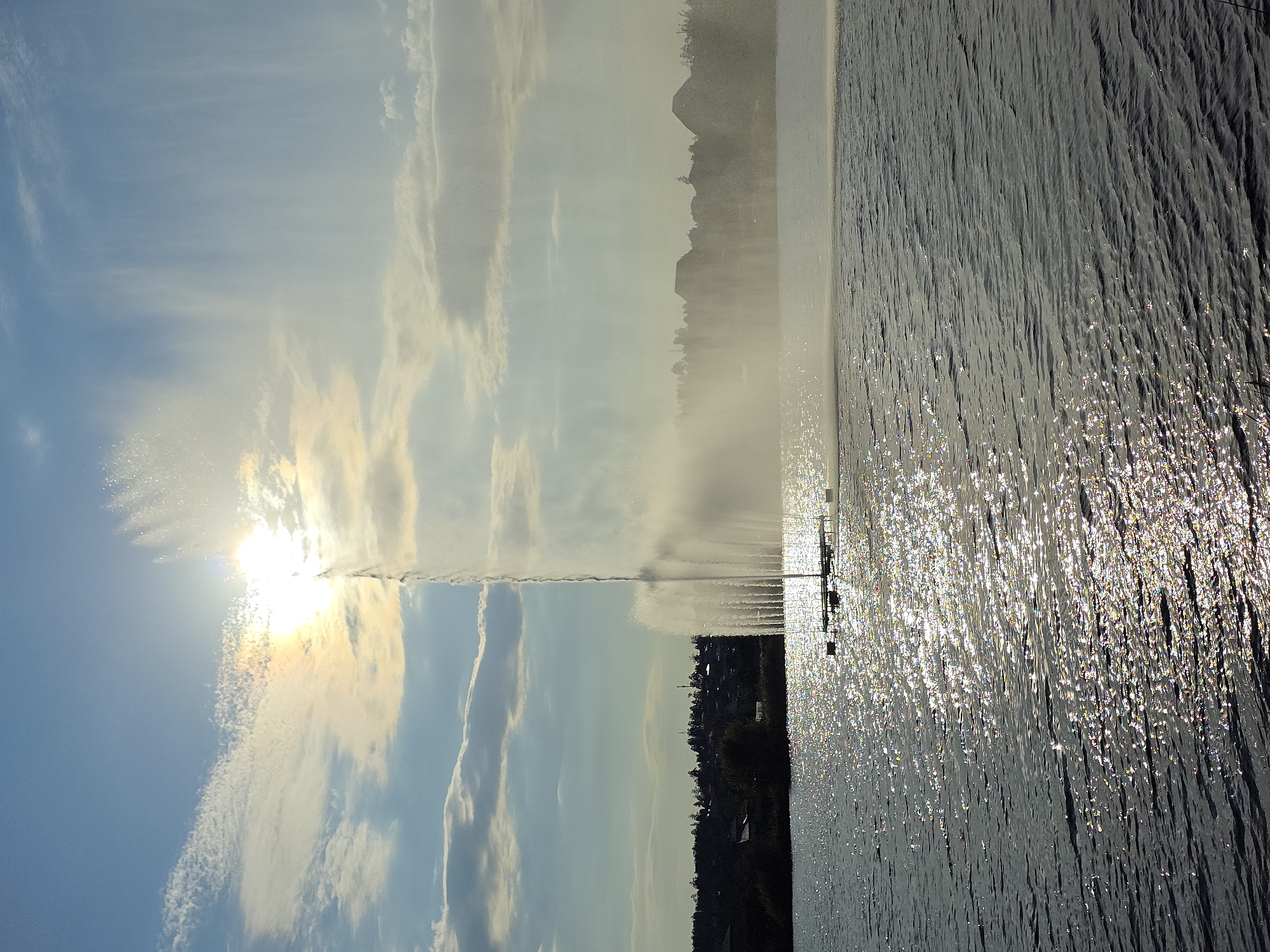 This image depicts a large fountain actively spraying water high into the air on a body of water, possibly a lake or reservoir, during the daytime. The sun is partially obscured by clouds, casting a bright glare on the water's surface, which is rippled and reflects sunlight. The background features a distant shoreline with a line of trees and buildings, suggesting a populated area. The sky is blue with scattered clouds, some of which are illuminated by the sun. The overall scene conveys a sense of open space and natural beauty, possibly a park or recreational area. This fountain is located on the Turgoyak Lake in the city of Miass, Russia.