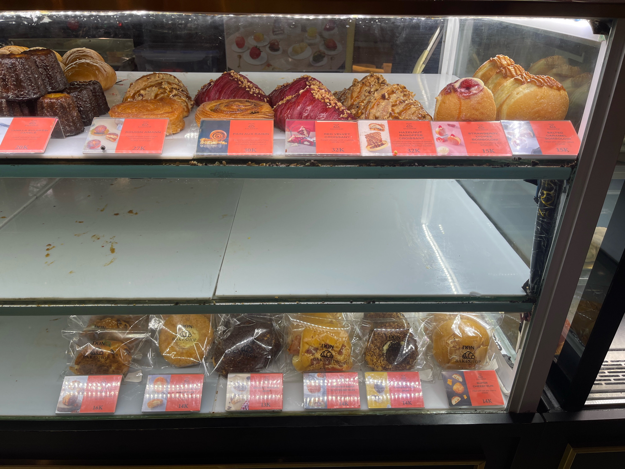 This is a medium-angle, eye-level, close-up, medium shot of a display case filled with baked goods at "DON Bakeshop," located in South Jakarta, Indonesia. The display case is well-lit and made of glass.
In the top shelf of the display case, various pastries are arranged. From left to right, there are canele, croissants, and what appear to be brioche or sweet rolls. Each pastry has a small card in front of it with its name and price. For example, the canele is labeled "VANILLA SULAWESI CANELE 20K," a pastry next to it is labeled "KOUIGN AMANN 27K," and a red velvet croissant is labeled "RED VELVET CROISSANT! 32K." Further down the row, there is a "HAZELNUT BANANA CROISSANT 32K," and a "STRAWBERRY LYCHEE BONBON! 15K," and a "SALTED CARAMEL BONBON! 15K."
The middle shelf of the display case is mostly empty, revealing the clean, white surface beneath the upper shelf.
The bottom shelf contains more baked goods, most of which are individually packaged in clear plastic. These are labeled with cards as well. Some of the labels visible are "16K," "14K," and "13K." One visible item in packaging is labeled "SUPER CHEESY BUN 14K."
The background is blurred, but it appears to be the interior of a bakery. The overall impression is one of a well-stocked and organized pastry shop.