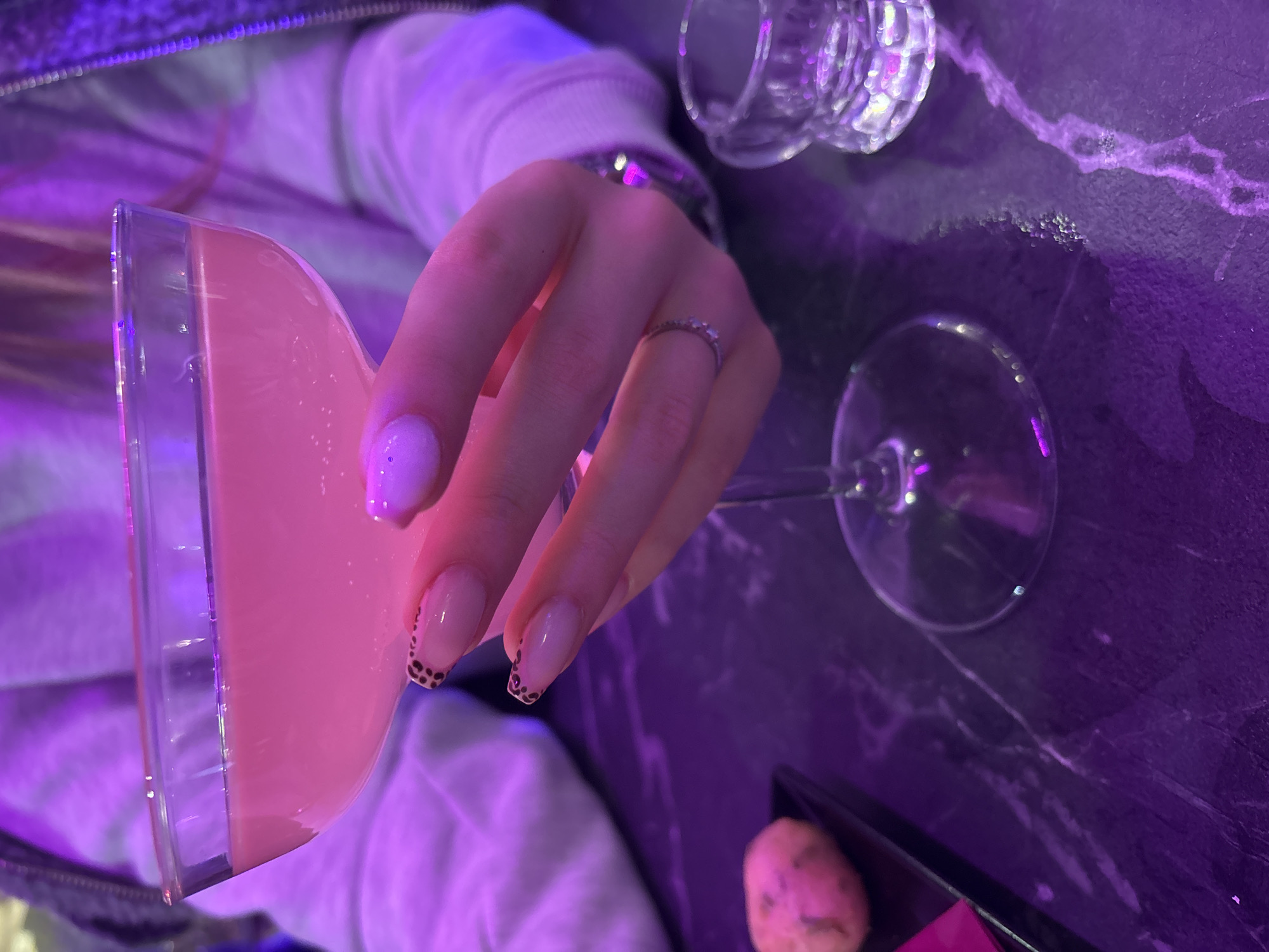 Here is a description of the image:
A woman is holding a pink cocktail in a wide-mouthed glass. She has a ring on her finger and an animal print design on the tips of two of her fingernails. Two other glasses, one empty and one with a short stem, are on the table. The table is black with a pattern resembling marble veining. A small black tray with a pink object and a pale sphere are in the corner of the image. The general lighting is tinted purple.
