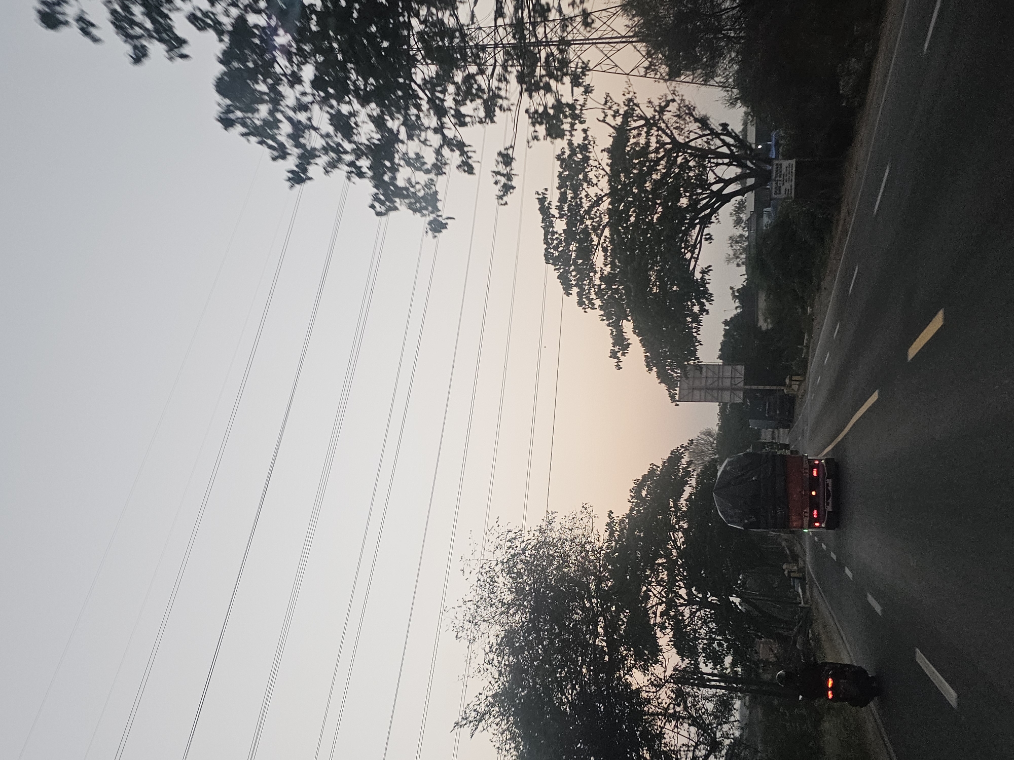 The image depicts a street scene in Jenu, Indonesia, during what appears to be dusk or dawn due to the soft, hazy lighting and the pale sky. A paved road with white and yellow lane markings stretches into the distance. In the foreground, a motorcycle is traveling on the left side of the road, its taillights illuminated. Ahead of the motorcycle, a red truck with a tarp-covered cargo bed is moving in the same direction. Further down the road, other vehicles, including what appears to be another truck, are visible. Dense foliage and trees line both sides of the road, with some trees casting shadows. Overhead, power lines are strung across the sky. A large metal pylon, likely part of an electrical transmission tower, is also visible on the right side of the image. The overall atmosphere is calm and suggests a rural or semi-urban setting.