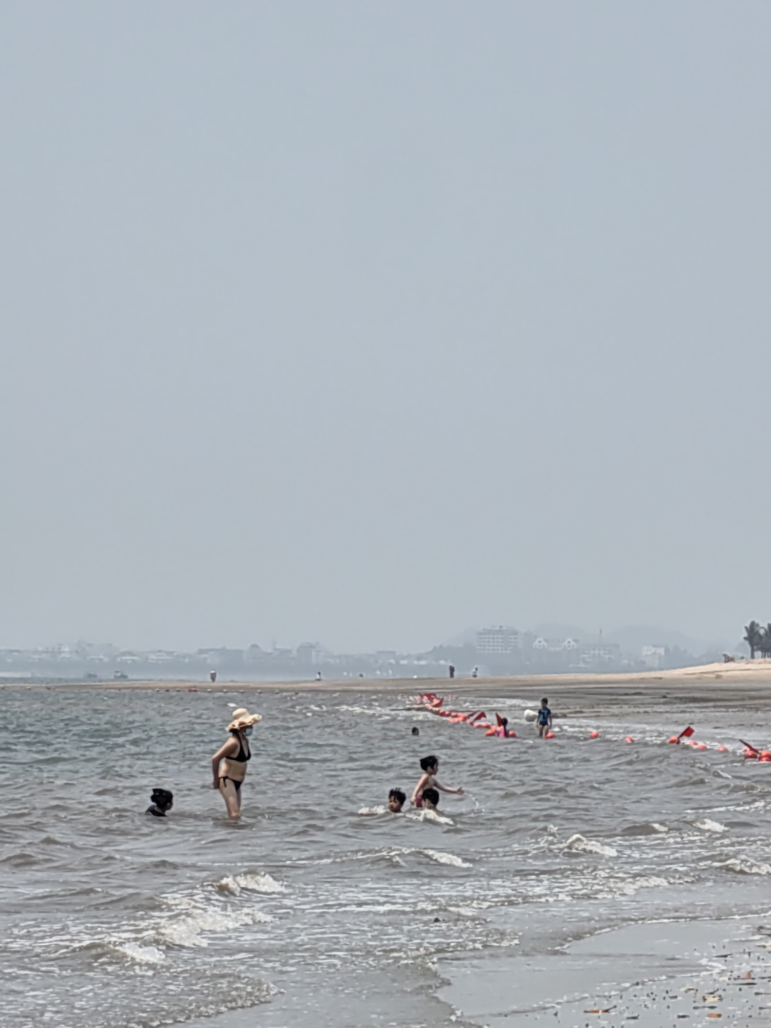 The scene depicts a beach on a hazy day. Several people are wading in the water near the shoreline, including a woman wearing a hat and bikini and a child with dark hair, and other children. Red buoys are strung together along the water, indicating a boundary. The beach itself is sandy, and in the background, there are buildings and trees, indicating a location in Bai Chay Ward, Vietnam.