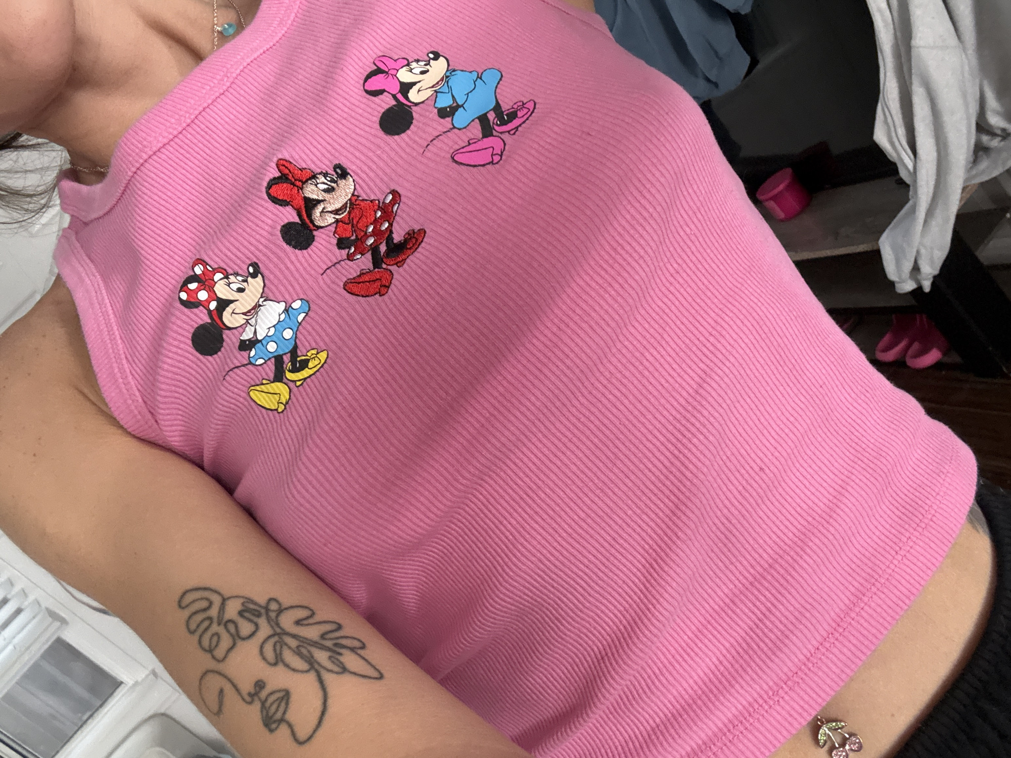 A person is wearing a pink ribbed tank top with three embroidered Minnie Mouse figures, one in blue, one in red, and one in blue. They have a tattoo of a face and leaves on their right upper arm. A cherry charm dangles from their waistline. The background includes clothing on a shelf, and a view of a window. The location appears to be indoors. There is no immediate indication that the location is in Seal Beach, California.