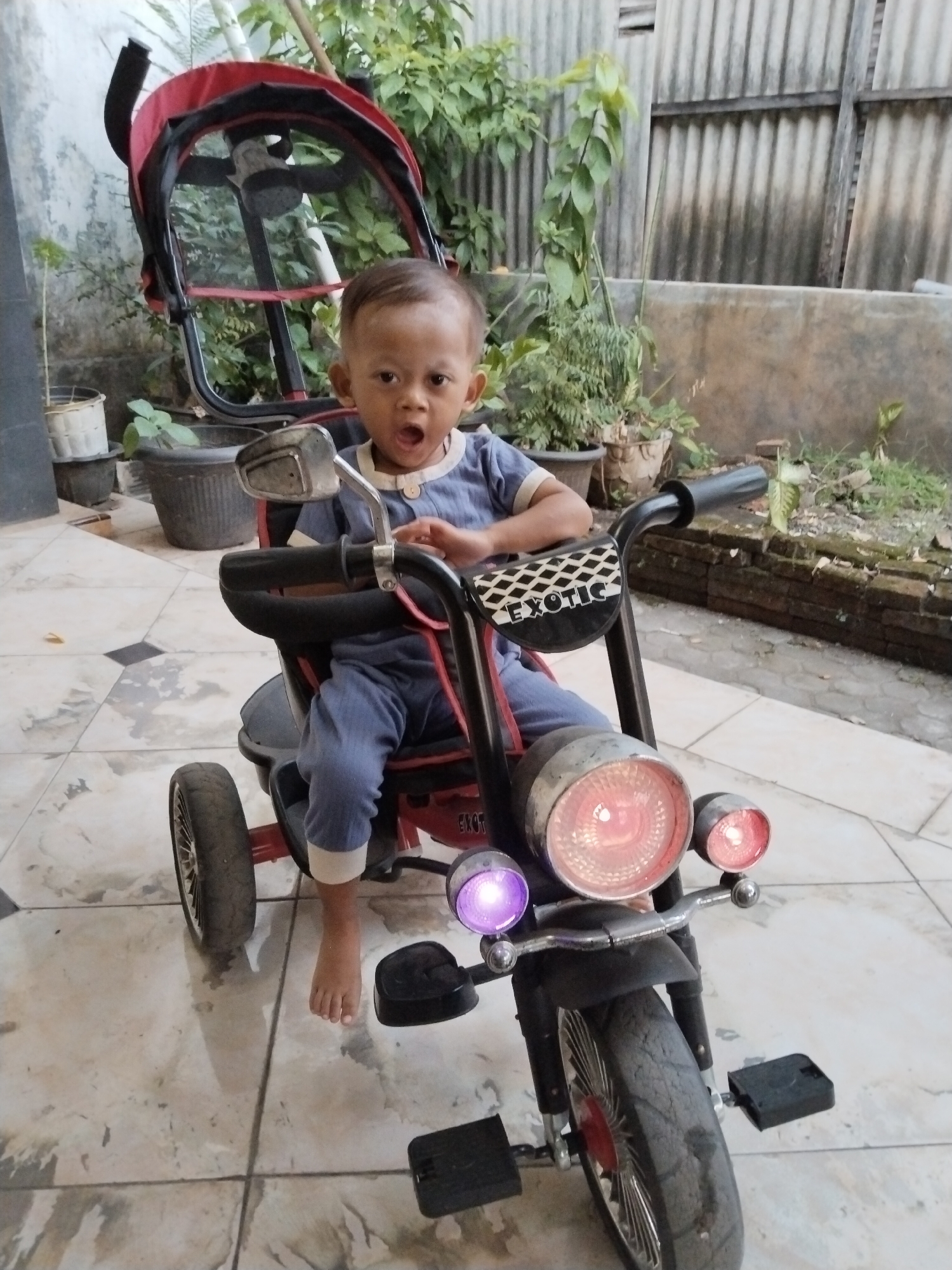A young child, appearing to be under two years old, sits in a red and black stroller tricycle with a handlebar that resembles a motorcycle. The child is wearing a blue onesie with white trim and has an open mouth, as if speaking or surprised. The tricycle has large front headlights, one of which emits a purple light, and two smaller red lights. The word "EXOTIC" is visible on a black panel on the tricycle's handlebars. The child's bare foot rests on the ground to the left. The tricycle is positioned on a tiled outdoor patio. In the background, there are various potted plants and a corrugated metal fence. The scene is likely in Kecamatan Pati, Indonesia, based on the context provided.