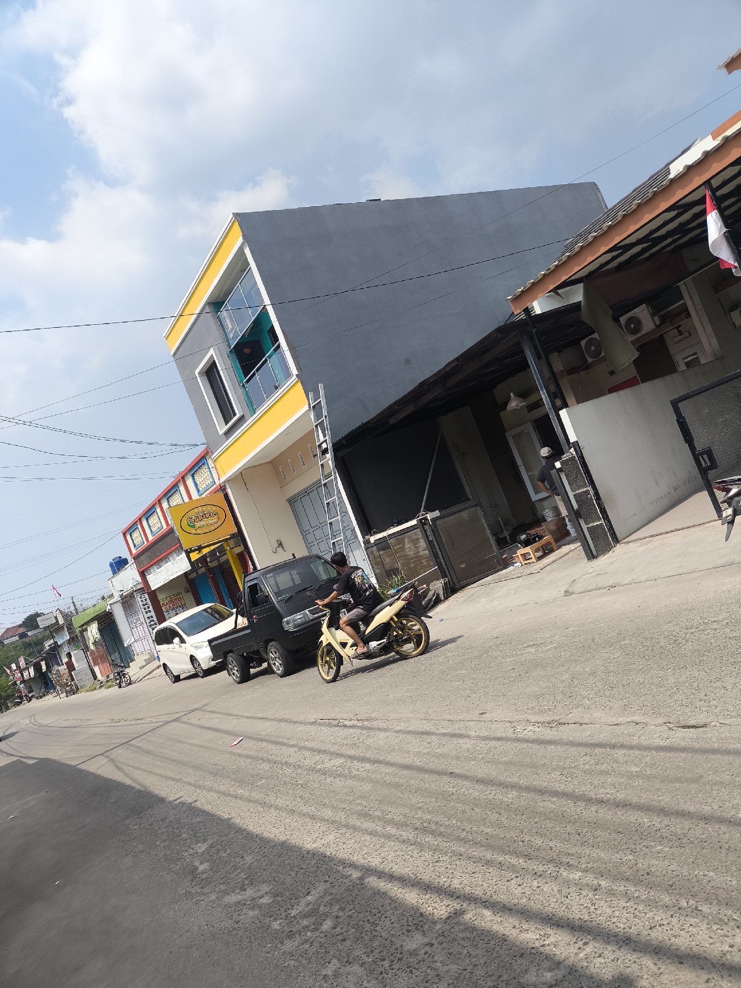 The image depicts a street scene in Cilegon, Indonesia, likely on a sunny day with scattered clouds. A two-story building with a gray and yellow facade dominates the left side of the frame, featuring multiple windows and a balcony. In front of this building, a black pickup truck is parked, and a person on a yellow motorcycle is moving past it. To the right of the pickup truck is a white van. Further down the street, other buildings and structures are visible, some with signs that are partially legible. An Indonesian flag can be seen on one of the buildings on the right. The road is paved, and shadows from overhead wires are cast across it. The overall impression is of a commercial or mixed-use area within a city.