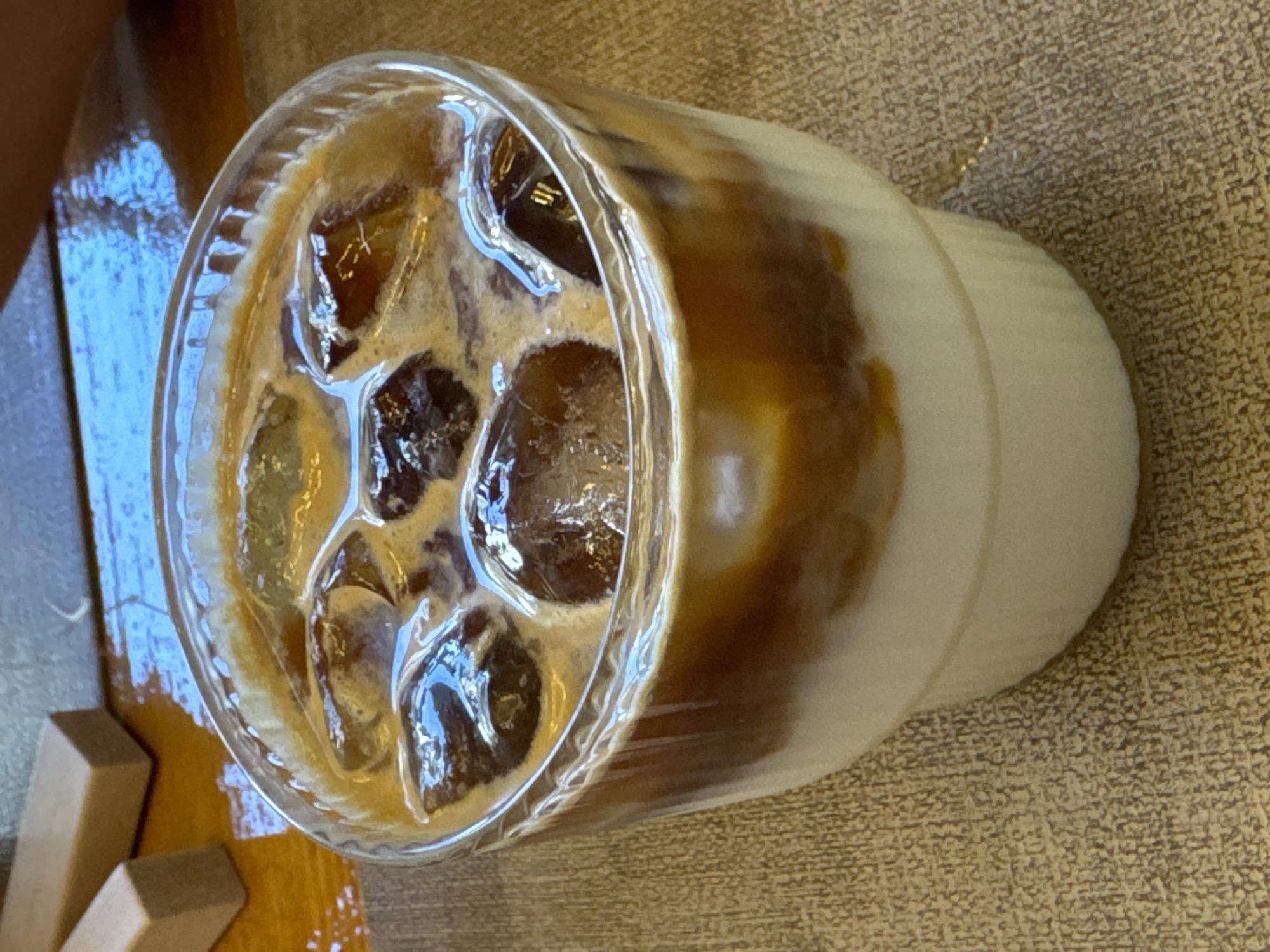 The image shows a close-up, high-angle view of an iced coffee drink in a clear glass cup. The coffee is layered, with a lighter cream or milk mixture on top of a darker coffee base, and ice cubes are visible within the drink. The glass has a ribbed texture on the lower half. The drink is placed on a textured surface that appears to be a light brown or beige tablecloth or placemat. To the left of the cup, a wooden surface is visible with two small wooden blocks. A hint of a person's hand or arm can be seen in the upper left corner of the image. The lighting suggests it is daytime, possibly indoors given the consistent lighting and the tablecloth. The scene appears to be a casual setting, possibly a cafe or a home. There is no visible text in the image.