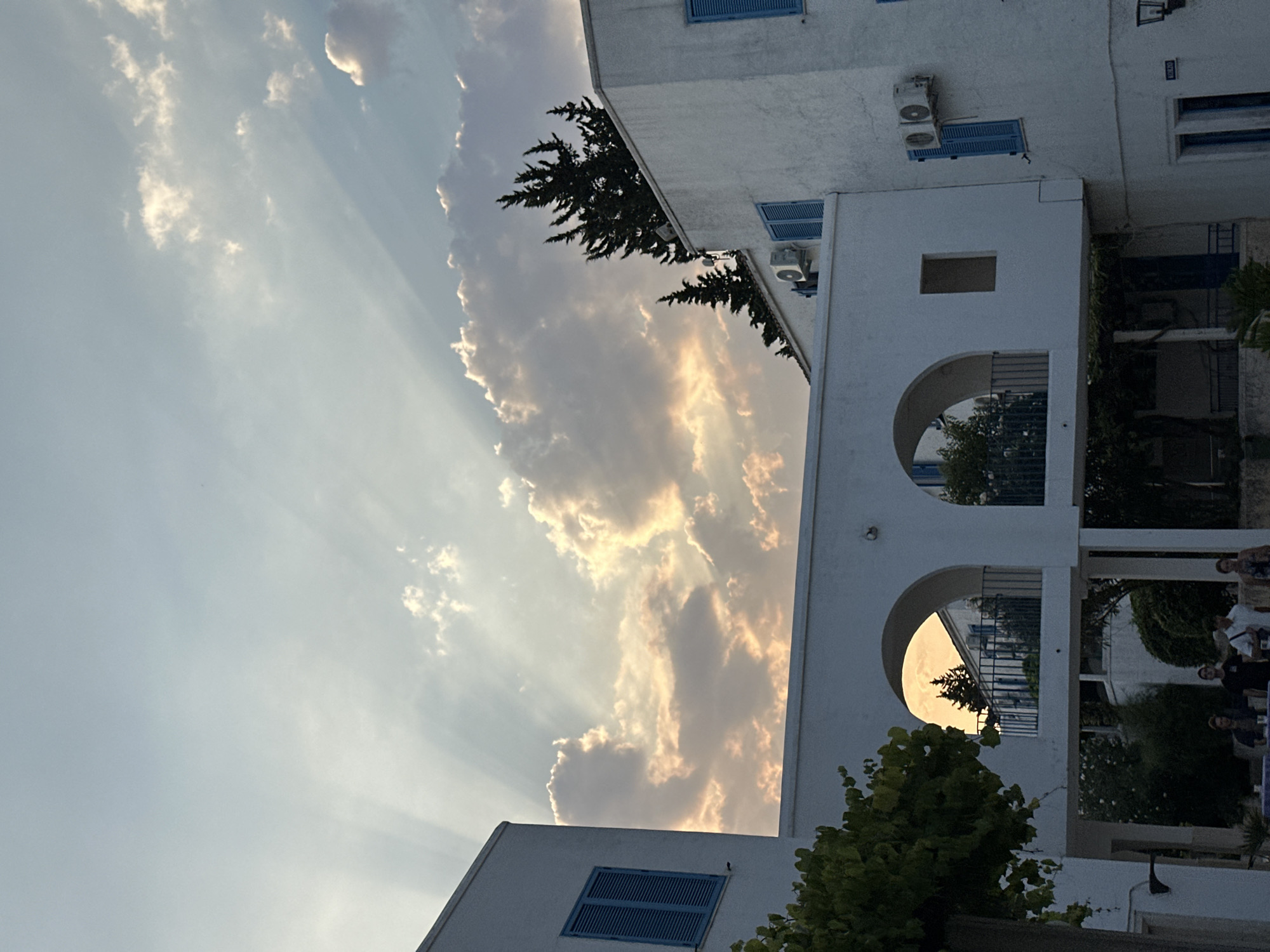 This image depicts a view of white buildings with blue shutters, characteristic of Mediterranean architecture, likely in Budva, Montenegro. The buildings are connected by an arched walkway. In the sky, crepuscular rays are visible, suggesting it is either sunrise or sunset. There are people visible in the foreground, some standing and conversing, others seated at tables. Trees and greenery are also present, adding to the outdoor setting. The overall scene conveys a peaceful and serene atmosphere.