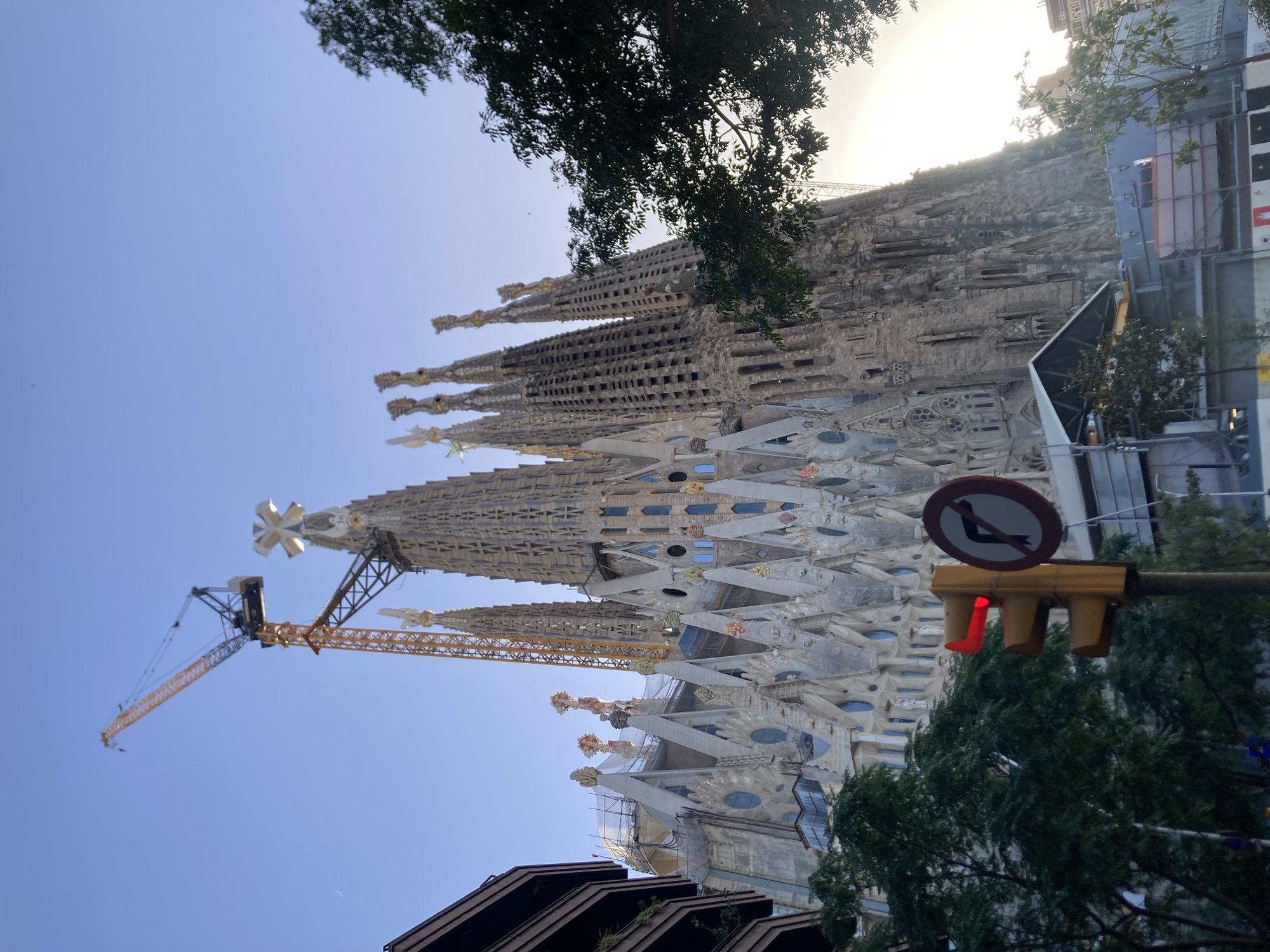 The image depicts the Sagrada Familia basilica in Barcelona, Spain, seen from a low angle against a clear blue sky. A tall yellow construction crane is prominently positioned to the left of the basilica, reaching above its spires. The basilica itself is characterized by its intricate, Gothic-inspired architecture, with numerous spires and detailed facades. Construction appears to be ongoing, with scaffolding visible on some parts of the structure. In the foreground, a traffic light displays a red light, and a circular traffic sign with a black arrow indicating a prohibited right turn is visible to its right. Lush green trees partially obscure the view of the basilica on the left and right sides of the image. The overall scene suggests an urban environment with ongoing construction work at a major landmark.