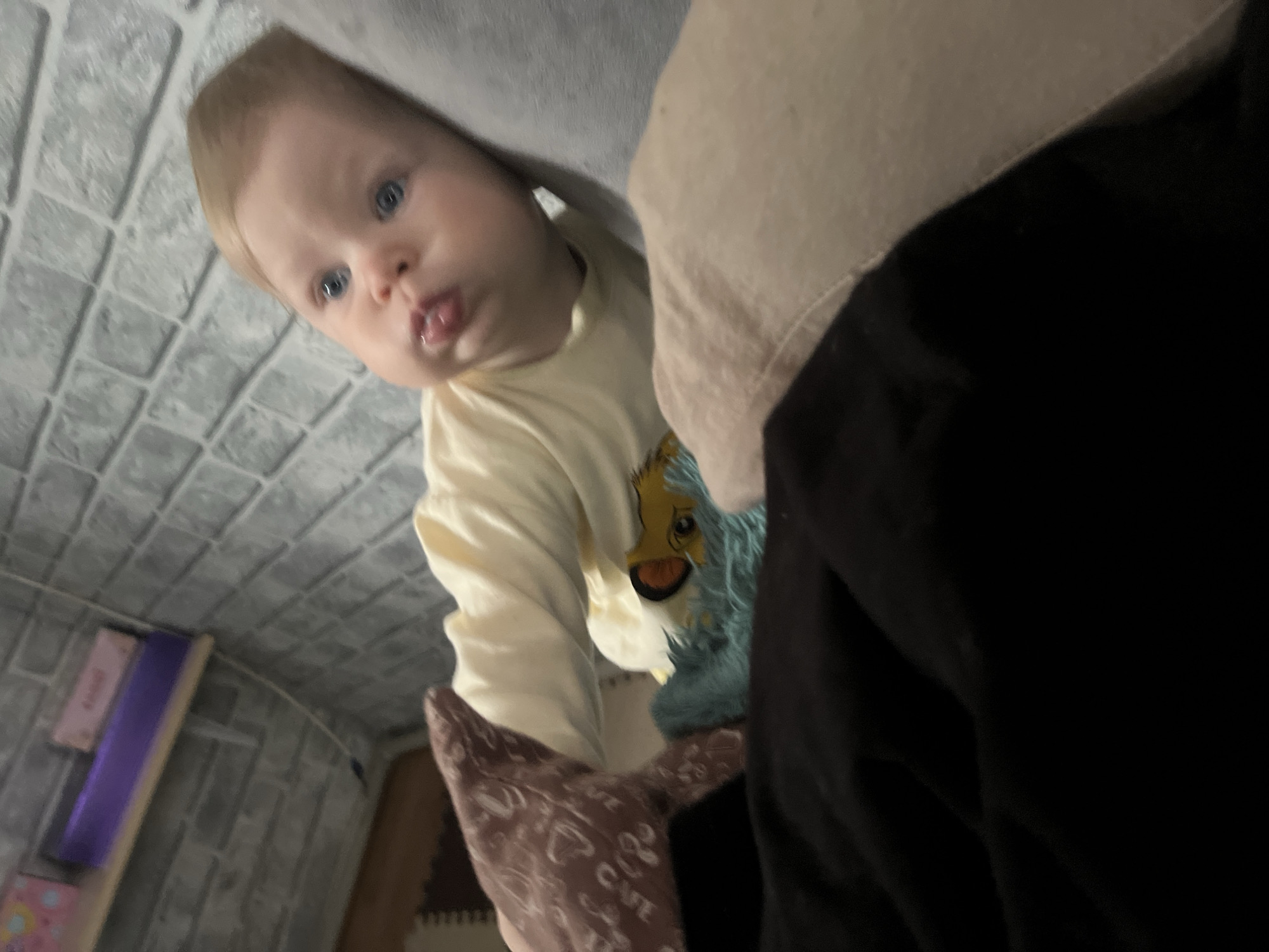 The image shows a baby with blue eyes and blonde hair wearing a yellow long-sleeved shirt with a colorful graphic. The baby is partially obscured by a white and black piece of clothing. The background appears to be a gray faux brick wall. The location is unknown, and there is no indication of Vesyoloye, Russia.