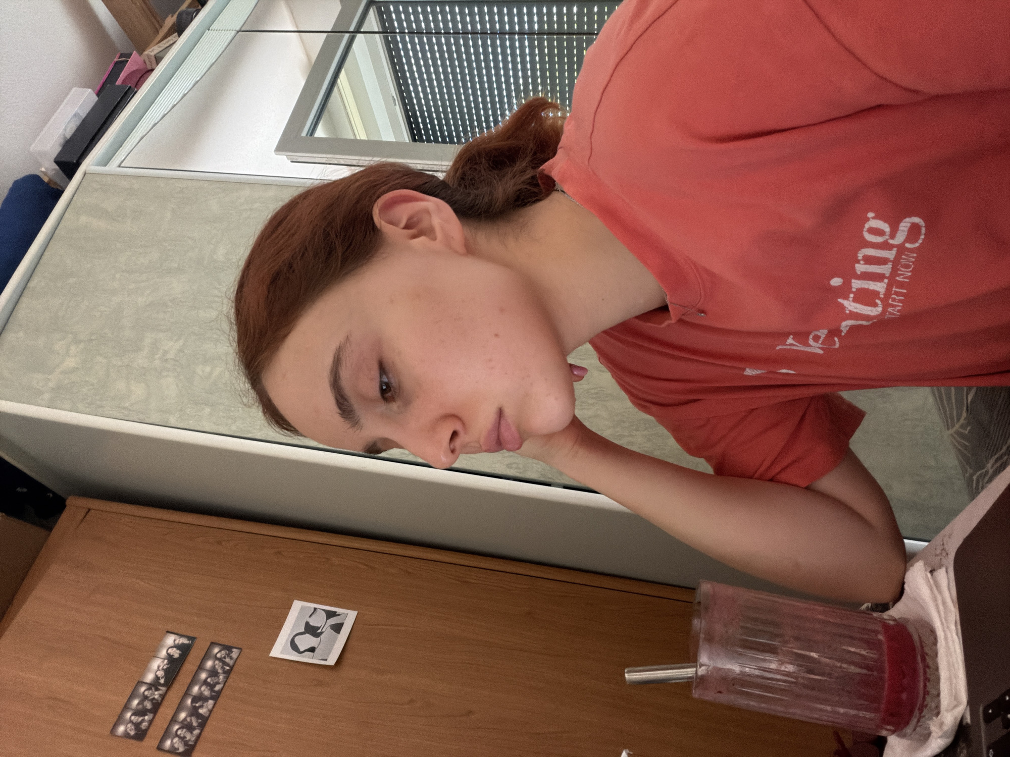 A young woman is depicted indoors, likely in a room in Strasbourg, France, during bright daylight. She is facing towards the left of the frame, with her head slightly tilted downwards and her left hand resting gently under her chin, suggesting a pensive or contemplative mood. Her hair is reddish-brown, pulled back from her face, and her fair skin shows light freckles across her cheeks and nose.
She is wearing a coral-colored or orange t-shirt. Text is visible on her shirt, partially reading "verting" with "ART NOW" below it, possibly part of a larger word like "Advertising." Behind her is a large window with venetian blinds, indicating an outdoor view and the time of day.
In front of her is a light-colored wooden dresser or cabinet. On its surface, there are two strips of black-and-white photo booth-style portraits and a single square print featuring abstract black and white shapes. To her right, a clear blender cup filled with a red liquid, possibly a smoothie, along with a metal straw, sits on a white cloth or surface. In the background, past the window, additional shelving with various items can be faintly seen.