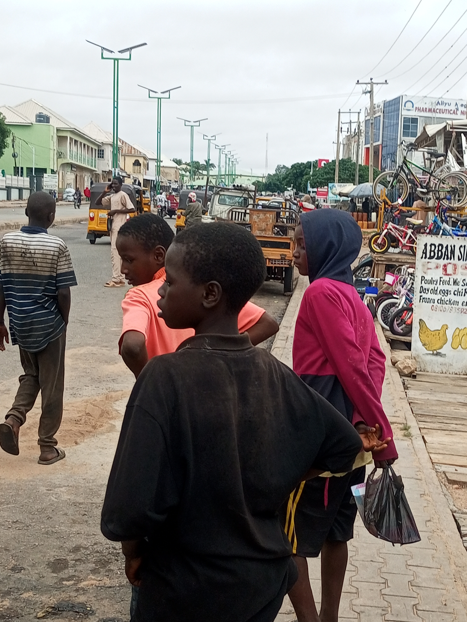 In Arawa, Nigeria, a street scene unfolds with a focus on a group of children. In the foreground, a boy in a black t-shirt is partially visible from the back. To his left, another boy, seen from the side, wears a bright orange t-shirt. Further left, a third boy walks away from the camera, wearing a striped t-shirt and dark pants. To the right of the orange-shirted boy, a child in a pink hooded sweatshirt holds a black plastic bag containing what appears to be money. The background reveals a bustling street with various vehicles, including yellow auto-rickshaws, a truck, and bicycles. Buildings line the street, some appearing to be shops with signage, including one for "ABBAN SIR." Overhead, streetlights with solar panels are visible, suggesting a modern infrastructure in this urban environment. The ground is a mix of asphalt and paved walkway.