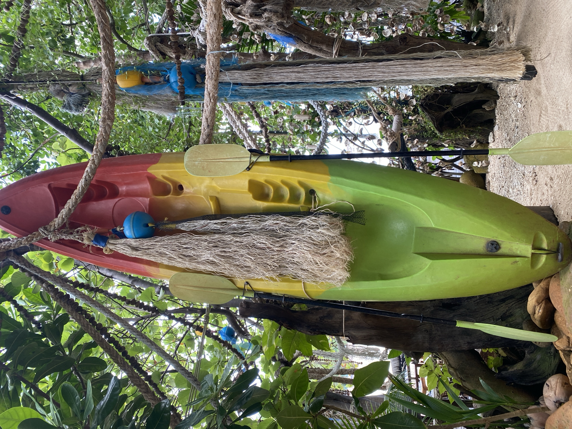 In ตำบลเกาะพะงัน, Thailand, a vibrant yellow and green kayak is suspended vertically from tree branches amidst lush foliage. Attached to the kayak is a coarse, light-colored net and a bright blue float. Two light-colored paddles with black shafts are propped against the kayak. The scene is further decorated with ropes and various strands of material hanging from the trees. The ground is covered in sand and small stones, with coconuts visible in the lower left corner. The environment suggests a coastal or tropical setting, possibly a resort or an outdoor recreational area.