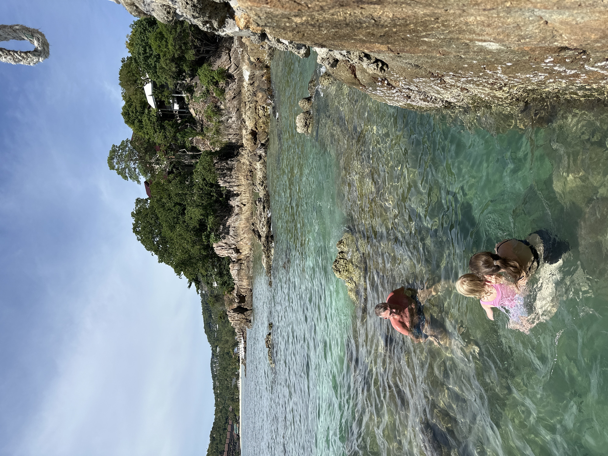 The image shows three people swimming in clear, shallow turquoise water near a rocky coastline. A man in the foreground is partially submerged, looking towards the camera. Behind him, two women are in the water, one holding a child. The scene is tropical, with lush green trees and rocky cliffs visible along the shore. Some white structures, possibly houses or resorts, are nestled amongst the trees on the hillside. The sun appears to be high in the sky, suggesting it is daytime, and the water is calm with gentle ripples. The overall mood is relaxed and peaceful, indicative of a vacation or leisure activity. A rope is visible in the top left corner of the image, suggesting a possible connection to a boat or dock. The location is identified as Koh Phangan, Thailand.