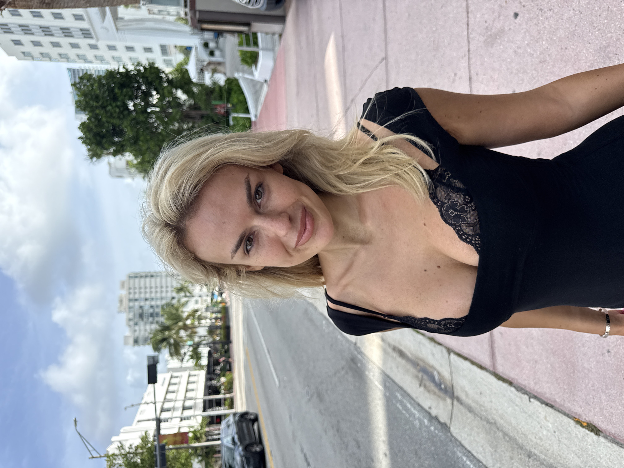 A blonde-haired woman is standing on a sidewalk in Miami Beach, Florida, United States of America, wearing a black dress with lace detailing. She is looking directly at the camera with a slight smile. The background reveals an urban environment with buildings, trees, and street traffic. A car is visible on the street, and the sky is partially cloudy. The sidewalk is pink and grey. The image appears to be taken at eye-level.