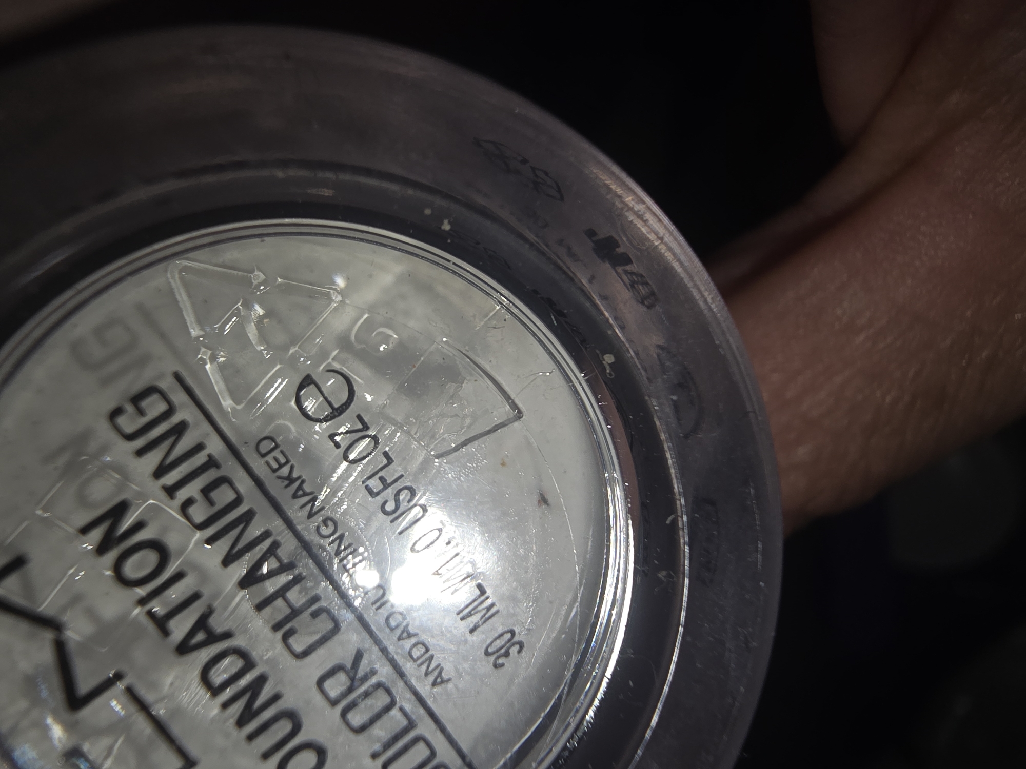 The image shows a close-up of a cosmetic product, likely foundation, in a clear plastic container. Text on the container reads "FOUNDATION COLOR CHANGING". Below this, it says "AND ADJUSTING NAKED" and "30 ML/1.0 US FLOZE". There is also a recycling symbol visible. The container is partially held by a person's fingers, which are visible at the bottom of the frame. The background is dark and out of focus. The image does not contain any information about the city of Ulan-Ude, Russia.
