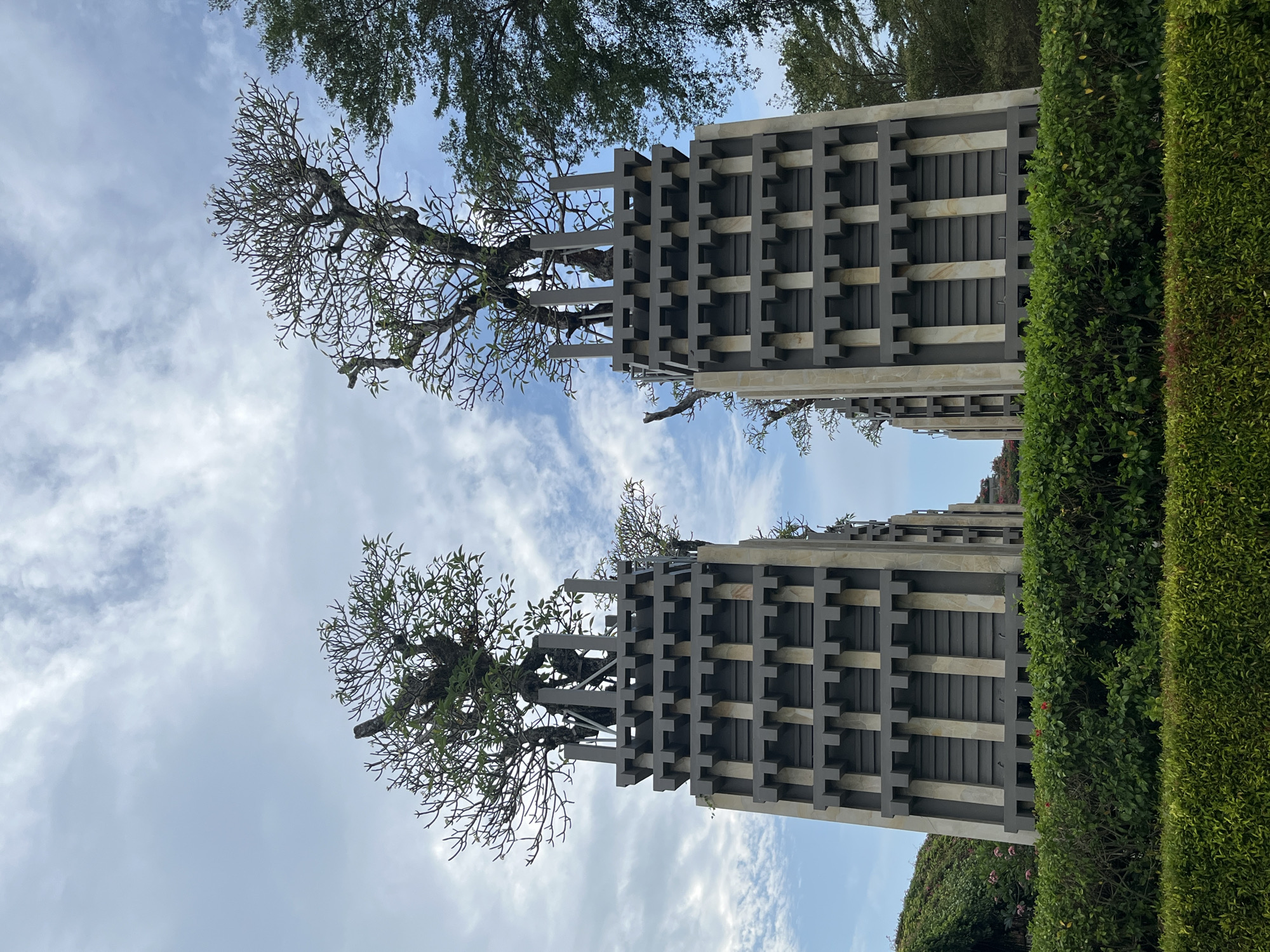 Two large, tiered, dark gray and beige rectangular structures stand in the foreground, topped with trees. The structures are detailed with a repeating pattern of recessed blocks and vertical pillars. The trees have sparse branches and a few green leaves, some of which appear to be pruned. In front of these structures is a neatly trimmed, dense green hedge. Behind the structures, the sky is partly cloudy with patches of blue. Further in the background, on the right side, lush green foliage from trees can be seen. The scene appears to be outdoors, in a well-maintained garden or park. Based on the architectural style and tropical vegetation, the location is likely a resort or public space in a warm climate. Given the provided location of Kuta Selatan, Indonesia, this image likely depicts a section of a hotel or resort within that area.