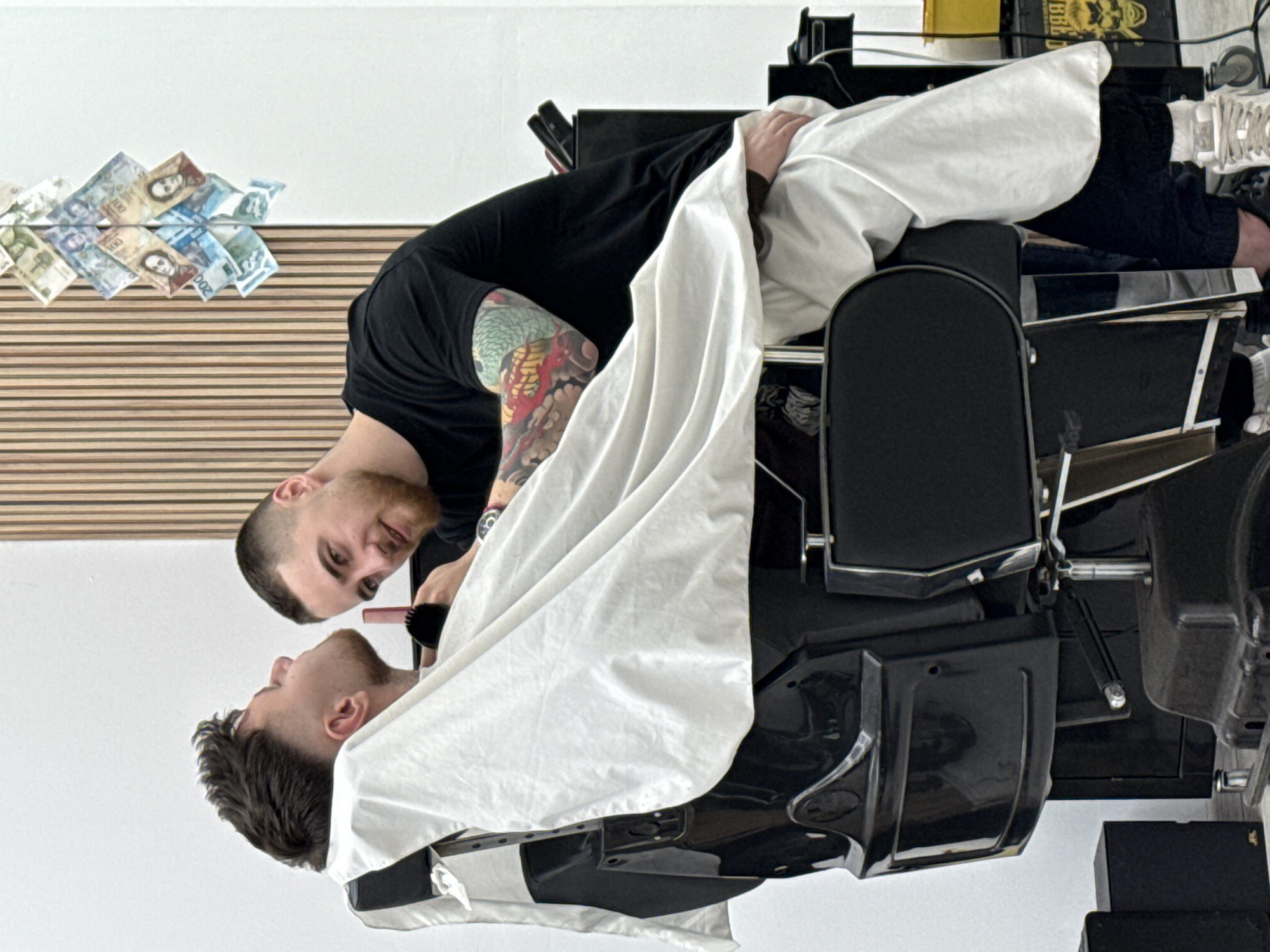 A barber in Sofia, Bulgaria, is giving a haircut to a client. The barber, wearing a black t-shirt and sporting a tattoo on his arm, leans over the client who is seated in a black barber chair and draped with a white cape. The barber holds a comb in his hand. Banknotes are displayed on the wall behind the barber.