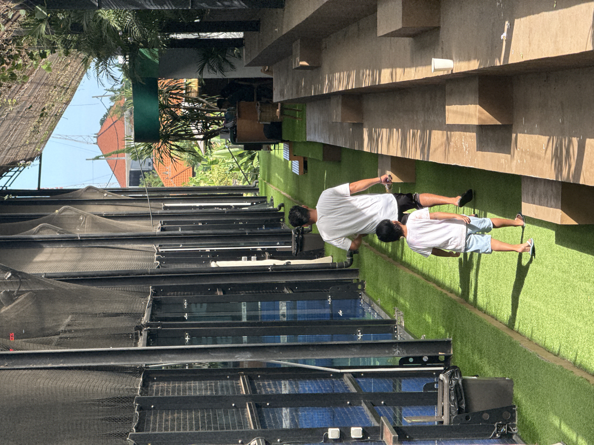 This is an outdoor scene in North Kuta, Indonesia, taken on a sunny day. Two people, an adult and a child, are standing on a bright green artificial turf lawn. The adult is wearing a white t-shirt and dark shorts, while the child is wearing a white t-shirt and light blue shorts. The adult holds a white string in their right hand. The lawn is bordered by a multi-tiered architectural structure, and a series of rectangular boxes with netting covers. Beyond the structure, there are buildings, trees, and foliage visible. The sky is blue.