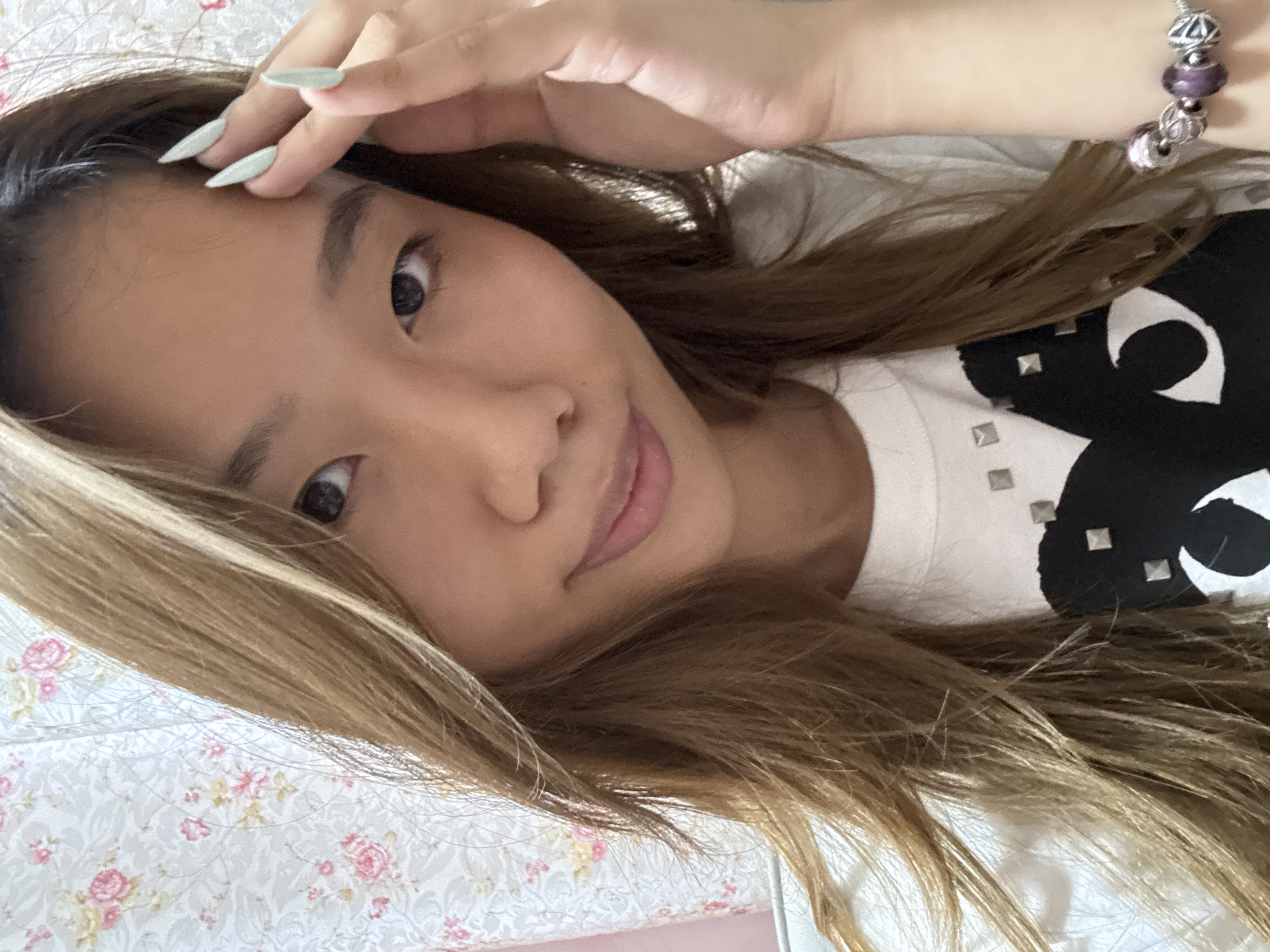 A young Asian woman lies on a floral patterned bedspread. She rests her right hand on her forehead. She is wearing a white t-shirt with a black design and silver studs. On her right wrist, she wears a silver bracelet with purple beads. She has long brown hair with blonde highlights. The photo was likely taken in a residential setting, but the specific location in Shapingba District, China cannot be determined from the image.