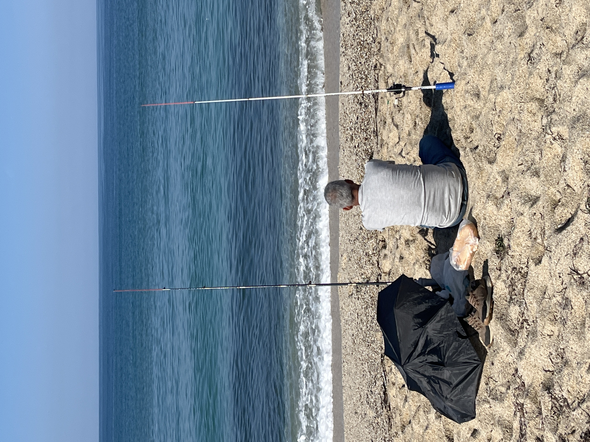 A man is fishing on a beach. He is sitting on the sand near the shoreline, with two fishing rods cast into the sea. An open black umbrella is nearby. The location is the beach in Alanya, Turkey. The man is wearing a gray shirt and blue jeans. The sea is calm and blue. The sky is clear.