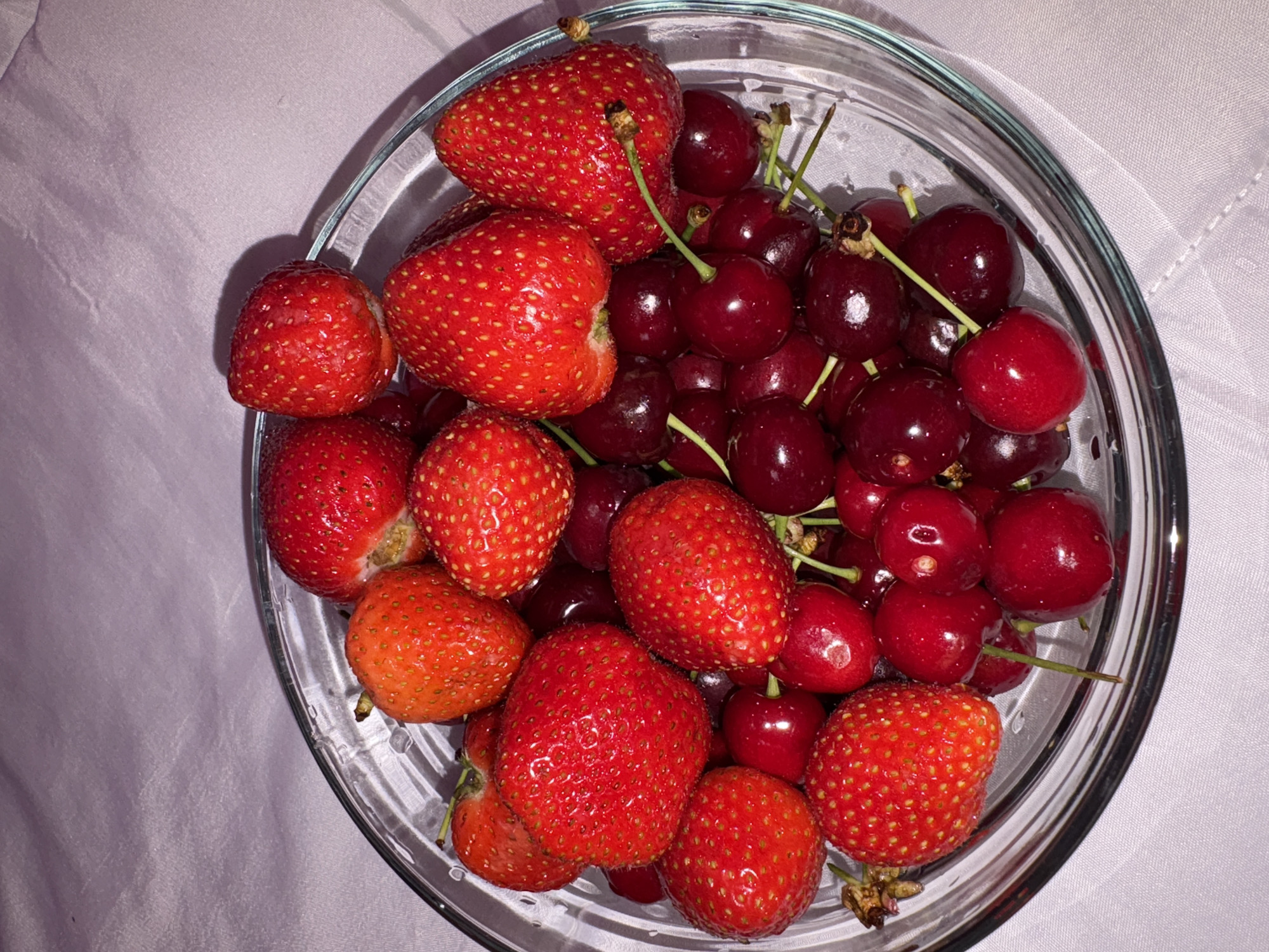 A clear, round glass bowl is centrally positioned, filled with an assortment of red fruit. The primary fruits are ripe strawberries and dark red cherries. Approximately two dozen strawberries are visible, characterized by their bright red color, textured surface with numerous small achenes, and occasional remnants of green calyxes or stems. These vary in size and orientation within the bowl. Interspersed among the strawberries are numerous dark red to maroon cherries, many retaining short green or brown stems. The cherries exhibit a glossy, reflective surface. The glass bowl itself has a visible rim and reflects ambient light. The bowl rests on a light-colored, possibly white or pale lavender, textile surface, which displays subtle folds and a slight texture, suggesting a tablecloth or fabric covering. No individuals are present. The specific geographical location, including the city of Vratsa, Bulgaria, cannot be determined from the visual content of this image.