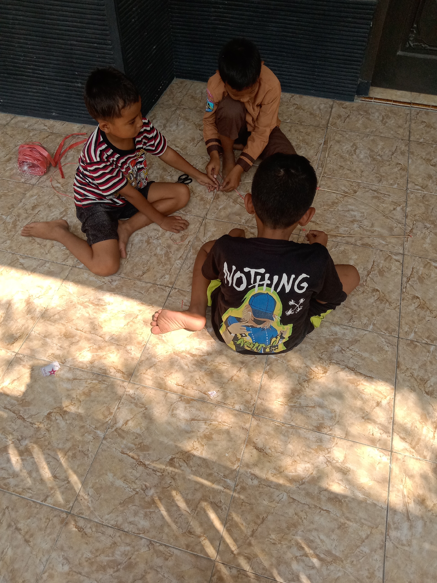 Three young boys are sitting on a tiled floor, engaged in an activity. The boy on the left, wearing a striped t-shirt and dark shorts, is looking down and holding scissors, with a spool of red string nearby. The boy in the middle, dressed in a tan uniform, is also looking down and appears to be working with thin sticks and string. The boy on the right, seen from the back, is wearing a black t-shirt with the word "NOTHING" printed in white and a graphic image on the back, along with dark shorts. The tiled floor is a light brown with a marble-like pattern. Sunlight casts shadows across the ground. The background shows a dark, slatted wall and a door. The scene is set outdoors, likely in a residential area in Stiaji, Indonesia, given the context provided.