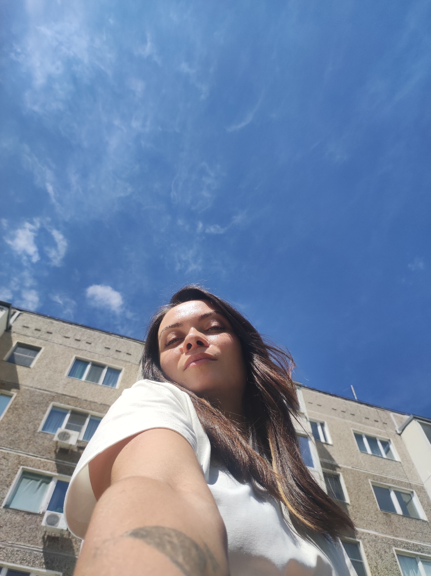 A low-angle, full-daylight shot captures a woman posing for a selfie against a backdrop of a blue sky with sparse cirrus clouds and a multi-story residential building. The woman, positioned in the foreground, is fair-skinned with long, dark hair highlighted with lighter strands. She wears a white t-shirt and has a tattoo visible on her extended arm. The building behind her is a typical urban apartment block, possibly located in Chaikovsky, Russia, given the architectural style. It is constructed with light-colored concrete, featuring symmetrically arranged windows, several of which have air conditioning units mounted below them.