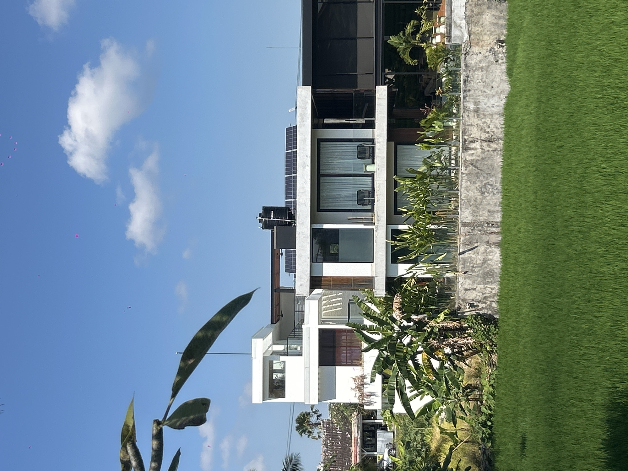 A wide-angle, eye-level, outdoor shot shows a modern, multi-story building with a dark roof and white facade in Mengwi, Indonesia, on a clear, sunny day. The building is situated behind a lush green rice paddy. The structure features large windows, some with white curtains, and a dark-tinted glass section. Solar panels are visible on the flat roof, along with a black water tank. Two chairs and a small table are visible on a balcony. Tropical foliage, including banana trees and other lush green plants, surrounds the lower levels of the building. The sky is blue with a few white clouds, and small pink objects, possibly flower petals or debris, are scattered across the sky. A leafy branch hangs from the top left of the frame.