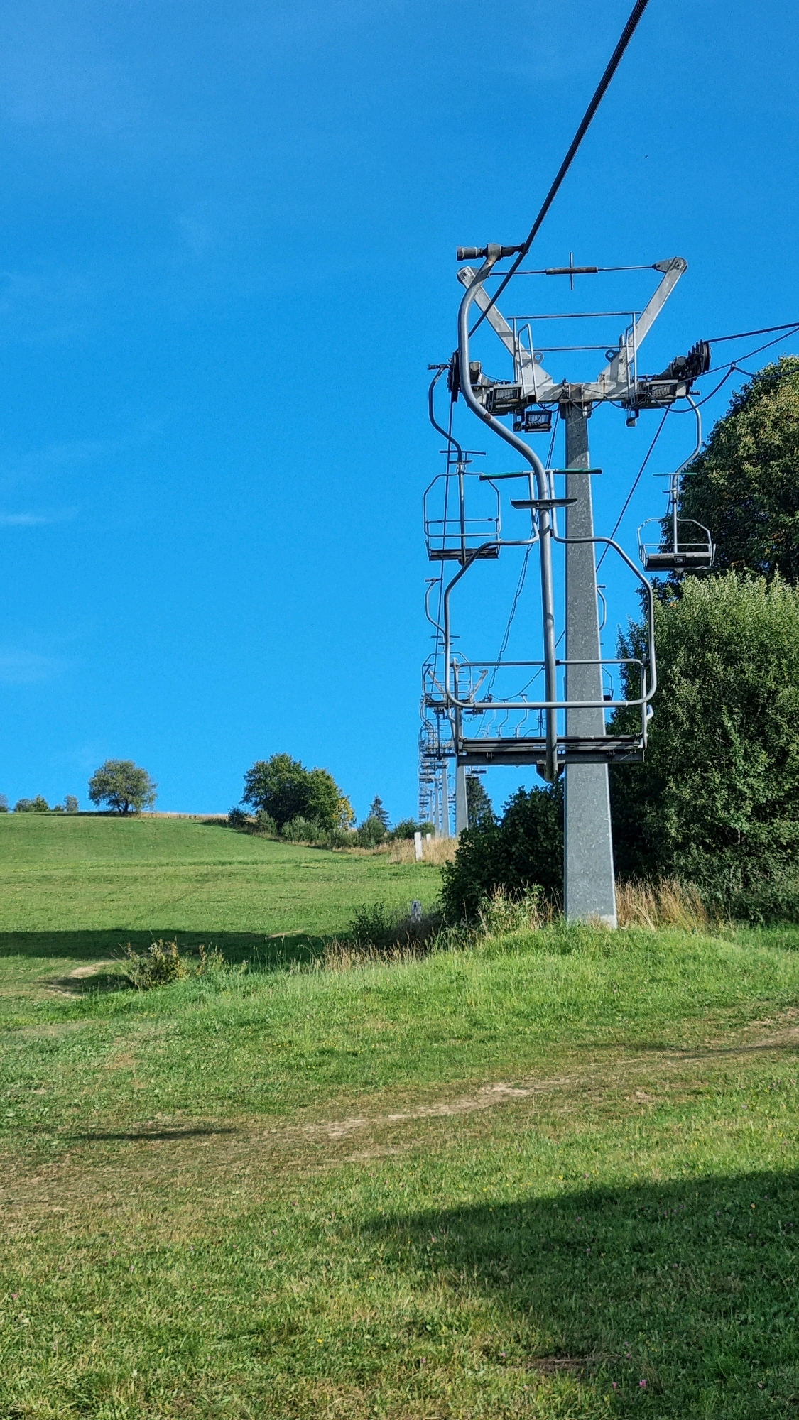 The image captures a ski lift on a bright, clear day in Izky, Ukraine. The ski lift, with its metal pole and suspended chairs, is prominently featured on the right side of the frame. The chairs are empty and hang from a thick cable that stretches across the top of the image. The ski lift is situated on a gently sloping green hillside, with lush grass and scattered trees. In the background, more trees and a clear blue sky dominate the scene. The overall impression is one of a serene, natural landscape, potentially in a rural or mountainous area.