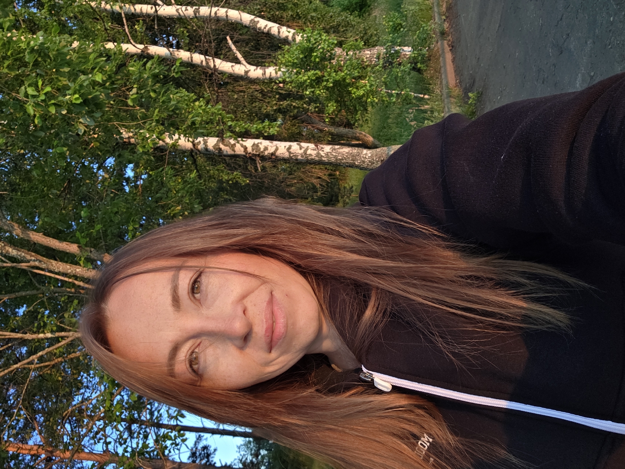 A medium shot captured outdoors features a woman with long, auburn hair and fair skin with freckles, looking directly at the camera. She is wearing a black hooded sweatshirt with a white zipper. The background consists of trees, with birch trees prominently displayed, and lush green foliage, suggesting a natural environment. The lighting indicates it may be late afternoon or early evening, with the sun casting a warm glow. The ground in the foreground appears to be dirt or gravel. The location is identified as Chaikovsky, Russia.
