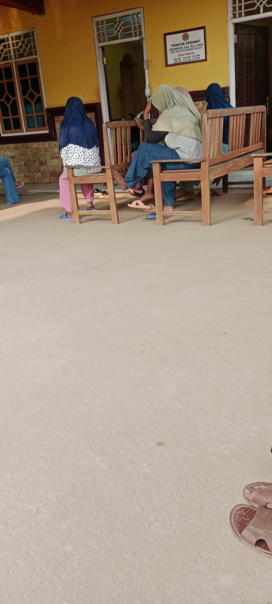 The image shows a waiting area outside a clinic in Gondang Rejo, Indonesia. Three individuals are seated on wooden benches. One person, wearing a blue hijab and a white and black polka dot dress, is seated on the far left bench. To their right, another individual wearing a light green hijab and blue jeans is seated, looking at their phone. A third individual, mostly obscured by the person in green, is visible to the right of them, also wearing a blue hijab. The clinic's sign is visible on the yellow wall, indicating "PRAKTIK PERAWAT" (Nurse's Practice) and listing operating hours. The ground is a light-colored concrete surface. In the foreground, a pair of brown leather sandals are visible. The overall scene depicts a calm, everyday setting at a local healthcare facility.