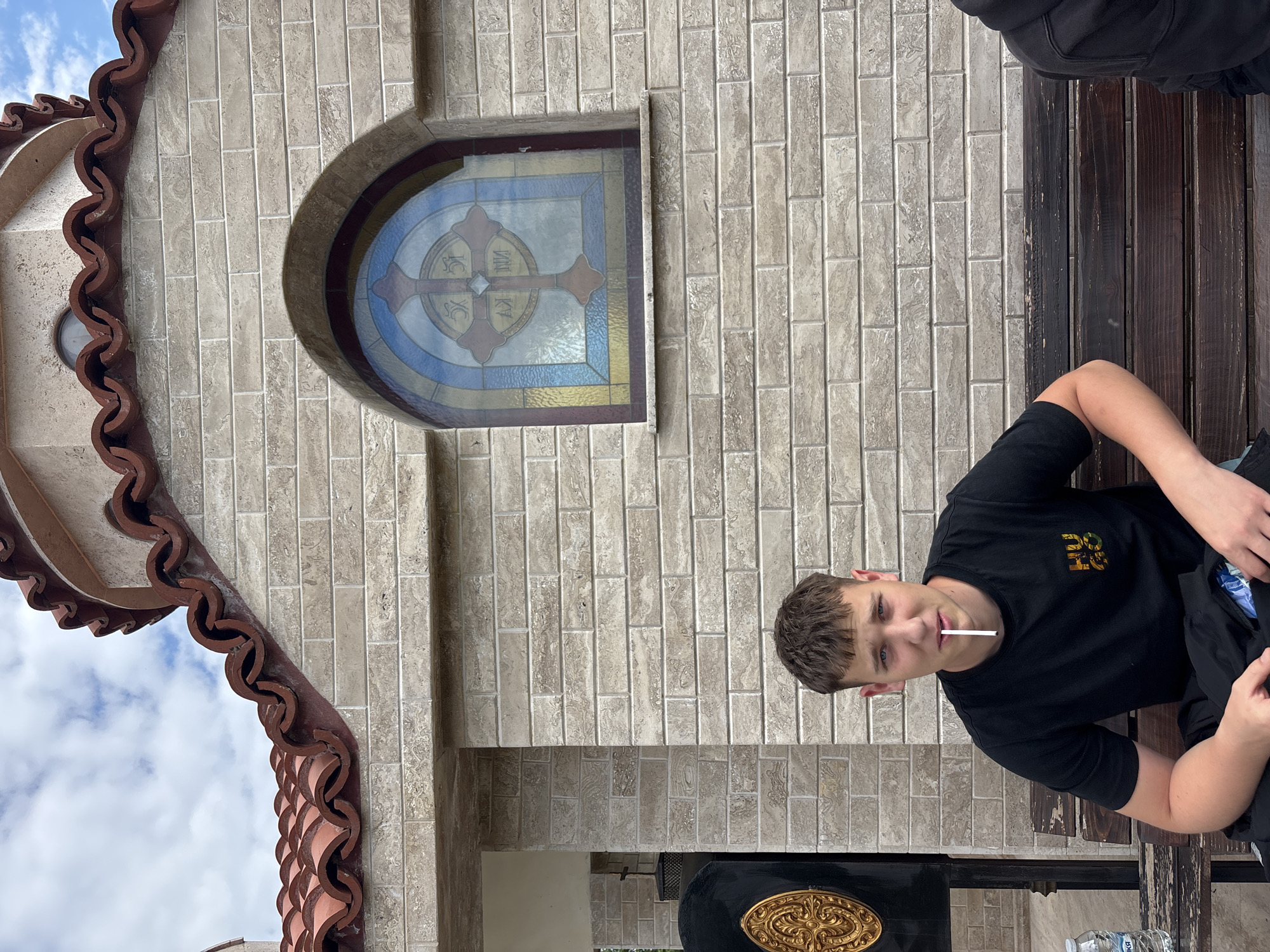 A young man sits on a wooden bench outside a building. The building has a tiled exterior with an arched window containing a stained glass cross. The man wears a black t-shirt and holds a thin, white object in his mouth. Part of the building's ornate roof and a decorative mailbox are visible. A water bottle is visible under the bench. The location is likely Zlatograd, Bulgaria due to the architectural style of the building, specifically the window and tiled exterior, which are typical of orthodox christian churches in Zlatograd.