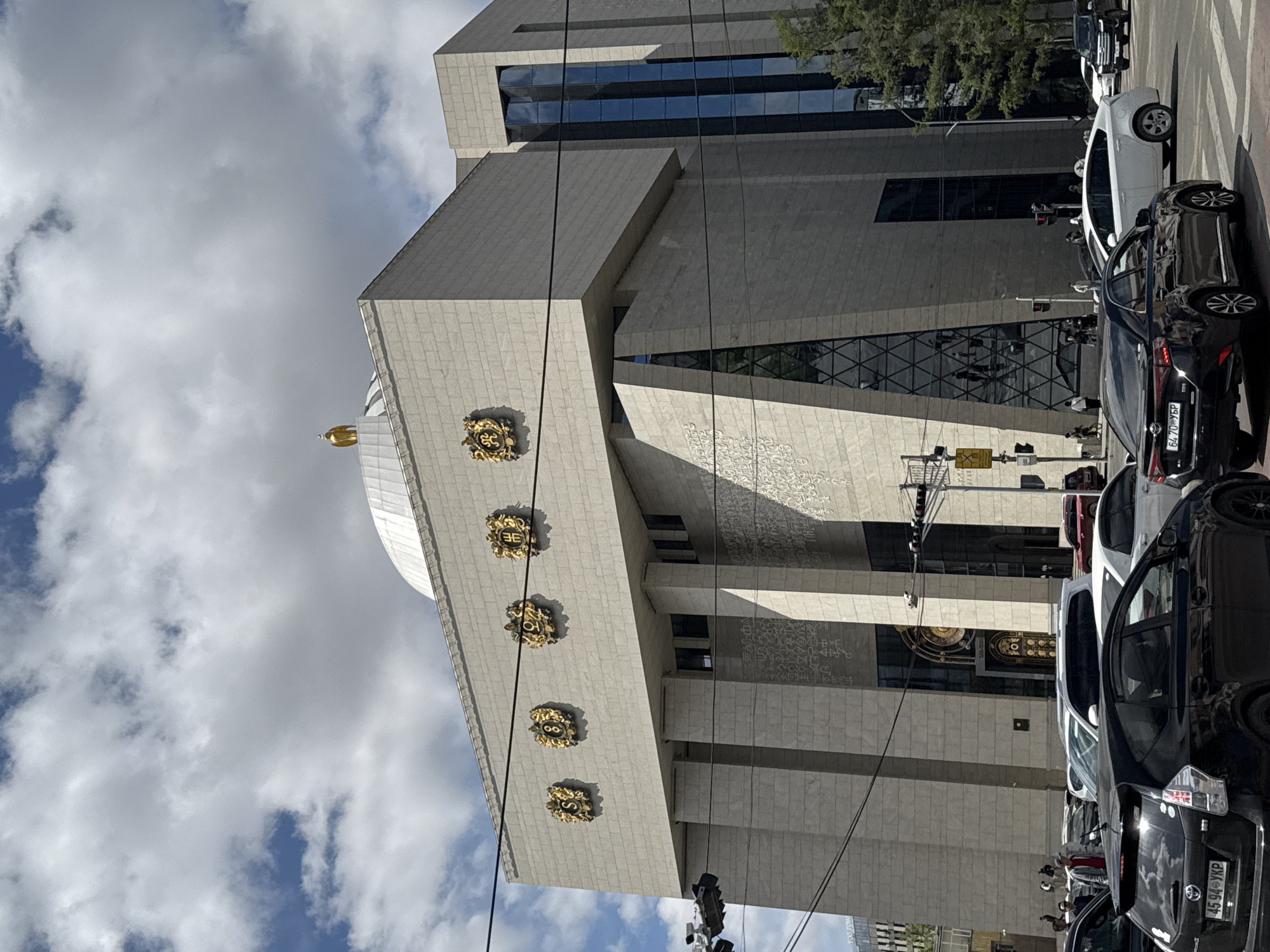 The image shows a section of a large, light-colored, multi-story building in Ulaanbaatar, Mongolia. The architecture features geometric angles and a dome topped with a golden spire. The facade is adorned with four prominent golden emblems. The sky is partially cloudy. The foreground shows traffic, including several parked cars and a traffic light. The location is identifiable as the State Great Khural (Parliament) building.