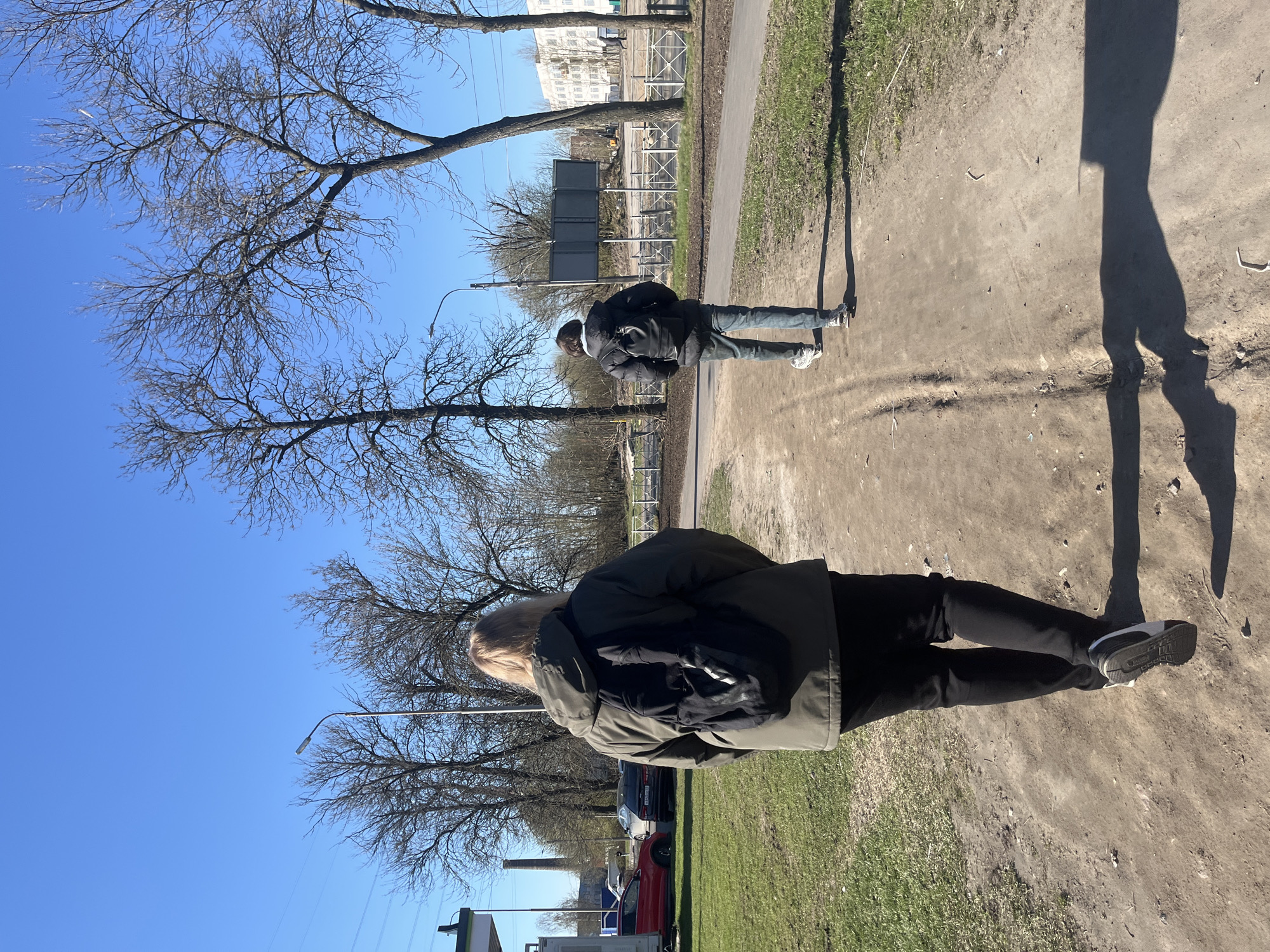 Here is a description of the image:
Two people walk on a dirt path adjacent to a paved path in what appears to be a park or green space. The sky is clear and blue, with bare trees visible overhead. The person in the foreground wears an olive-green jacket, black pants, and sneakers. They also have a black backpack. The second person in the background wears a dark jacket, blue jeans, and light-colored sneakers. They are walking away from the viewer on the paved pathway.
In the background, beyond the paved path, there is a fenced area and some buildings in the distance. There are a few cars visible in the lower left corner of the image.
The location is Saint Petersburg, Russia. There is a high probability based on the presence of architecture resembling Stalinist-era structures and the general ambiance consistent with the city. Further corroboration would require more specific landmarks.
