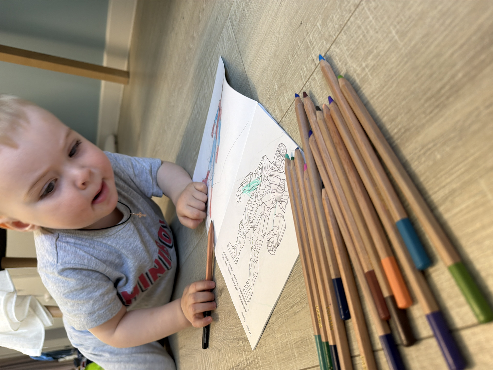 A toddler is on a light-colored wood-patterned floor coloring in a coloring book. The toddler is fair-skinned with blond hair, wearing a gray T-shirt with a red design and a black cord necklace with a cross pendant. The coloring book is open, showing a Marvel character on one page and a Marvel Spider-Man character on the other page. A collection of colored pencils with natural wood bodies and colored ends lies beside the coloring book. The child is holding a colored pencil. The location is unlikely Votkinsk, Russia.