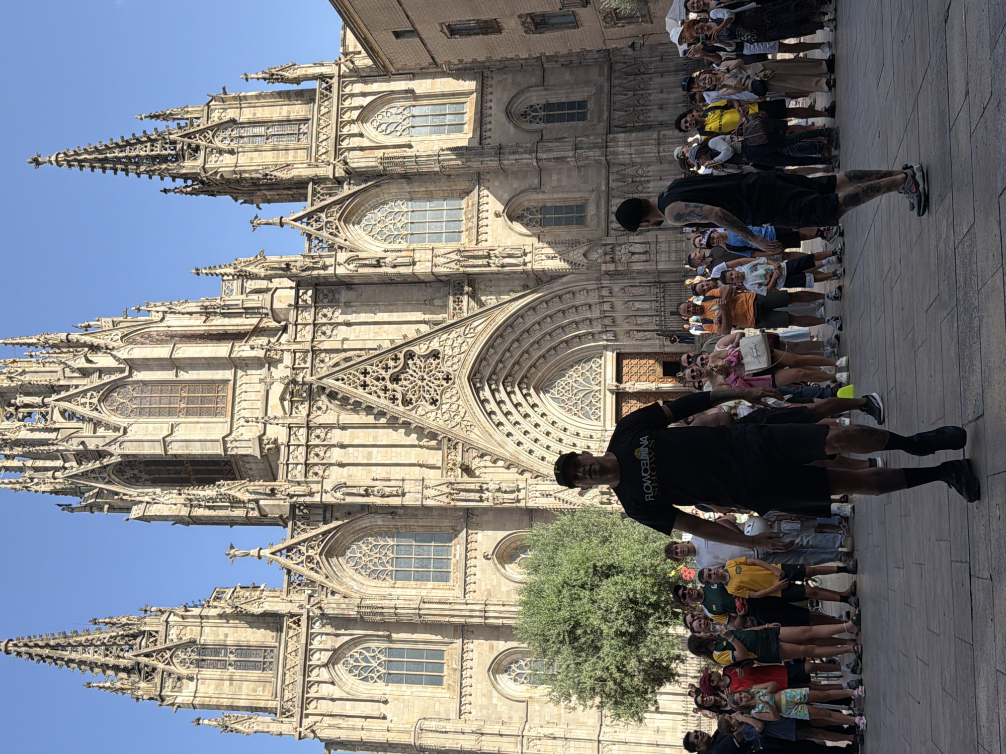 The image depicts a street performance in front of the Barcelona Cathedral, located in the Gothic Quarter of Barcelona, Spain. In the foreground, two male performers are visible. The performer on the left is wearing a black t-shirt with the logo "FLOWBARCELONA SHOWS", black shorts, black socks, and black shoes. He is facing the camera. The performer on the right is wearing a black tank top, black shorts, and athletic shoes. He is facing away from the camera, with his back to the viewer. Both performers have tattoos.
A crowd of spectators is gathered behind the performers, observing the performance. The spectators are diverse in age and gender, and many are holding up phones, presumably to take pictures or videos. They are casually dressed, suggesting a warm weather day.
The Barcelona Cathedral is a prominent Gothic-style structure dominating the background. Its facade features intricate architectural details, including spires, arched windows, and sculptures. The sky above is clear and blue. The ground in the foreground is paved with stone. The scene suggests a lively and engaging atmosphere in a historic urban setting.