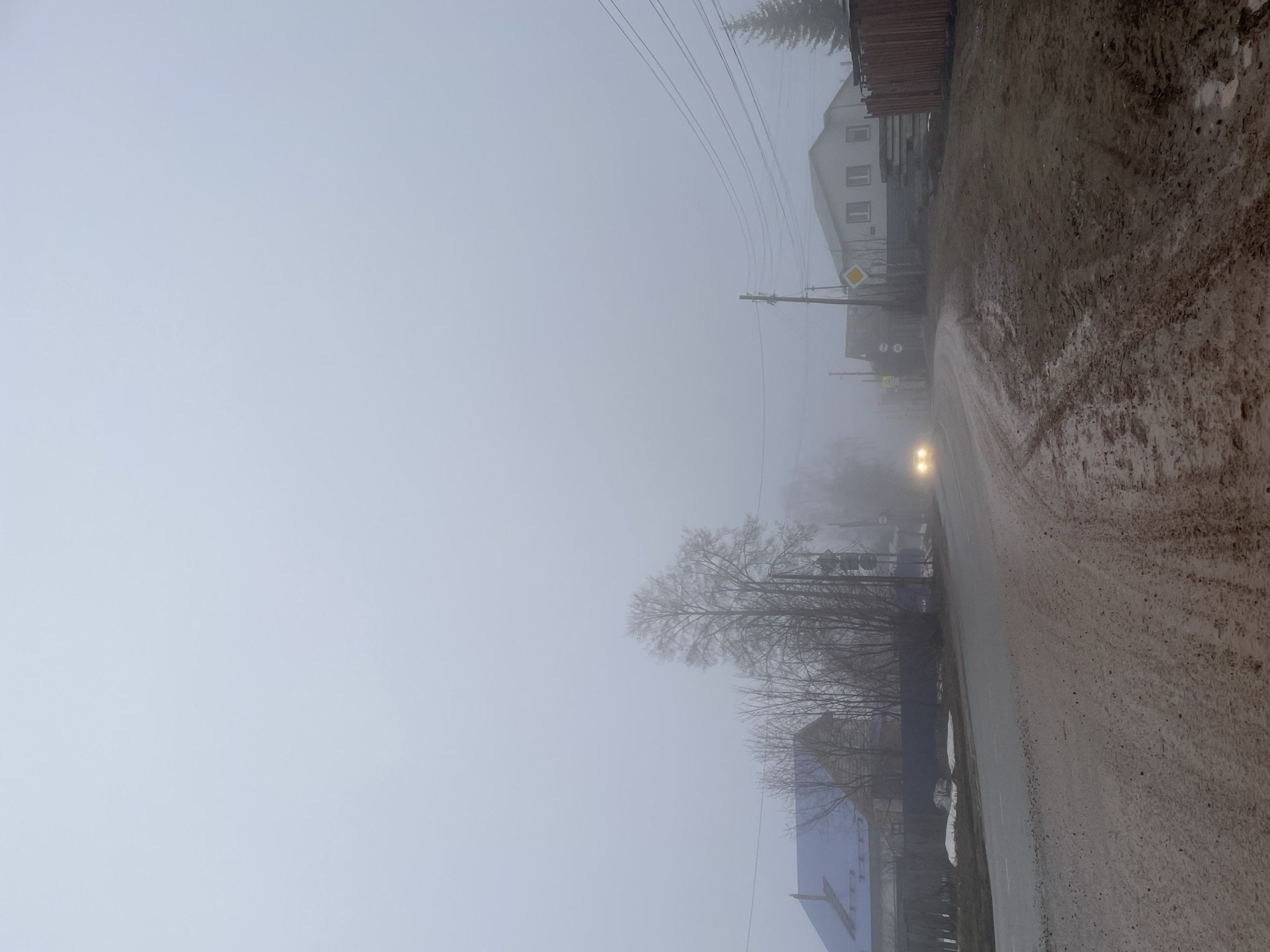 The image depicts a street in Фоки, Russia shrouded in heavy fog. The road is muddy and wet, with a car approaching in the distance, its headlights visible through the fog. Buildings line the right side of the street, partially obscured by the fog. Utility poles with overhead lines run alongside the road. Bare trees and a road sign are also visible, adding to the desolate atmosphere of the foggy scene.