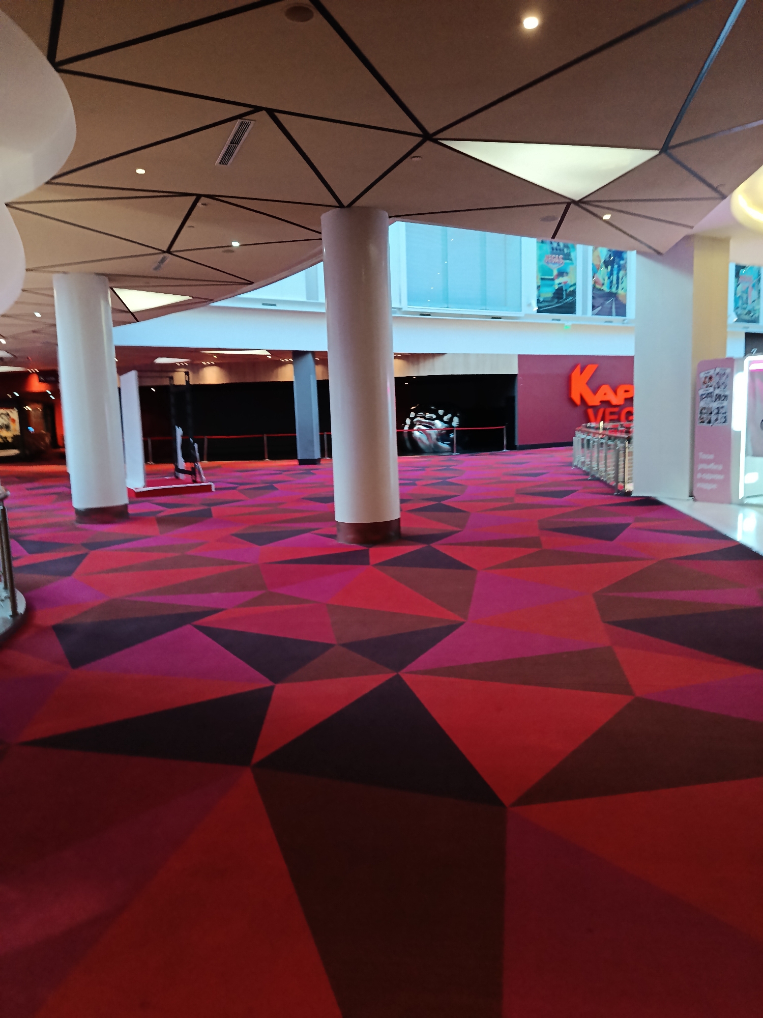 The image depicts the interior of what appears to be a modern entertainment venue, possibly a cinema or casino, with a striking geometric carpet in shades of red, pink, and dark purple. Large white cylindrical pillars support a geometrically patterned ceiling with black accents. In the background, a large sign with orange lettering reads "KAP VEG." To the left, a large, abstract silver sculpture is visible behind a red velvet rope. Further back, dark walls suggest entrances to rooms or auditoriums. There are no people clearly visible in the frame, but the scene suggests a public space. The overall aesthetic is contemporary and vibrant.