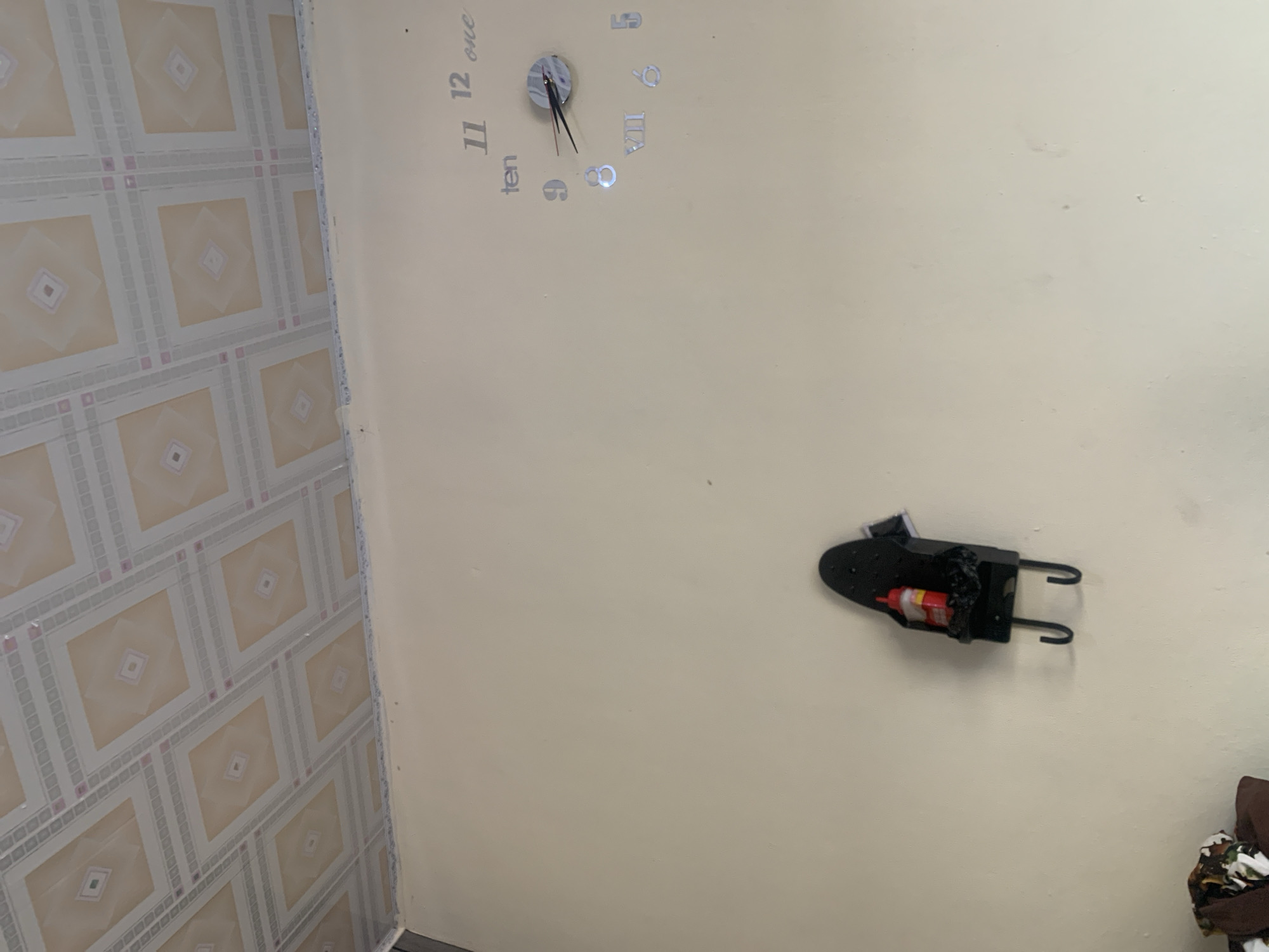 This image depicts an indoor scene, likely a wall in a room. A clock with Roman numerals and Arabic numerals is affixed to the wall. There is also a black wall-mounted organizer or shelf with two hooks, holding a red bottle with a yellow label. The wall is painted a light beige color, and the ceiling features a patterned design with squares. The image is taken from a low angle, looking upwards. The exact location in Jauro Yinu, Nigeria, is not specified within the image itself.