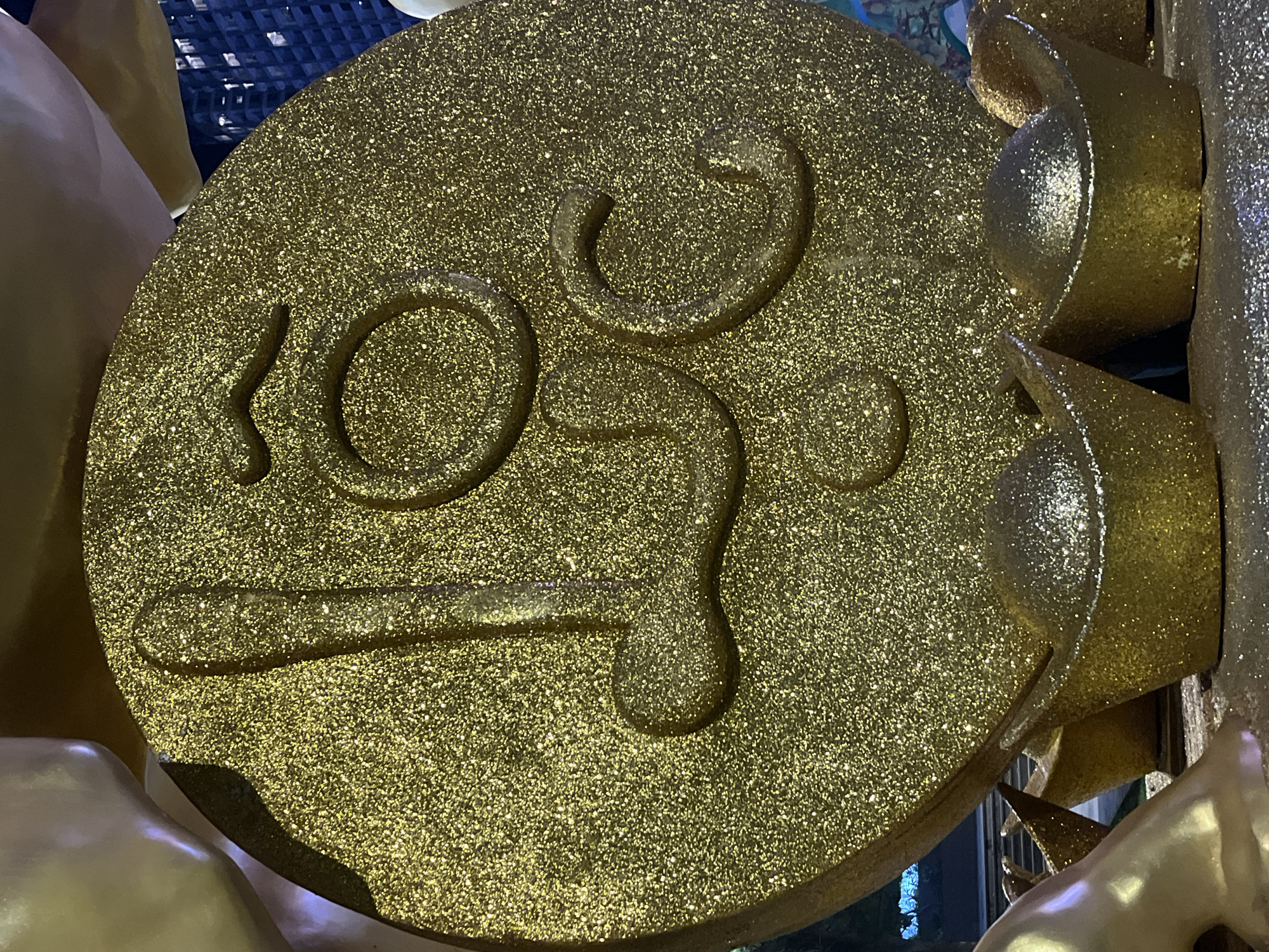 This is a close-up, eye-level photograph of a large, gold, glitter-covered coin-shaped decoration featuring raised Vietnamese text. The text appears to be "Tài Lộc", a common phrase associated with wealth and prosperity, often used during Lunar New Year celebrations. The coin is the primary subject, dominating the frame. The background is slightly out of focus, revealing hints of other decorative elements, possibly related to a festival or public display. In the foreground, several smaller gold, glitter-covered objects resembling Chinese gold ingots are visible, further reinforcing the theme of wealth and good fortune. The lighting is bright, emphasizing the glitter and golden hues of the decorations. The image does not show any people or specific urban landmarks that would definitively place it in Nha Trang, Vietnam, though the style of decoration is consistent with cultural celebrations in Vietnam.
