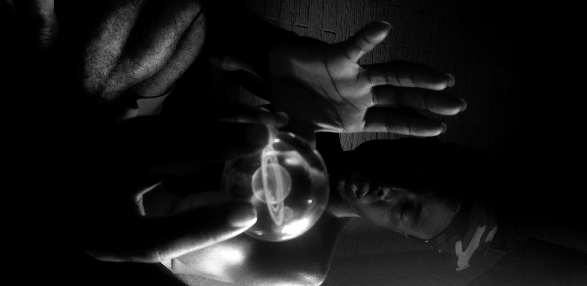 A black and white, low-light photograph depicts a person holding a lightbulb with an image of the planet Saturn inside, in front of their face. The person is likely male, with short hair and a camo headband. Their chest and arms are muscular. One hand holds the base of the lightbulb, while the other hand is extended above it. The person is shirtless. The background shows a dark, textured wall. Location is unknown.