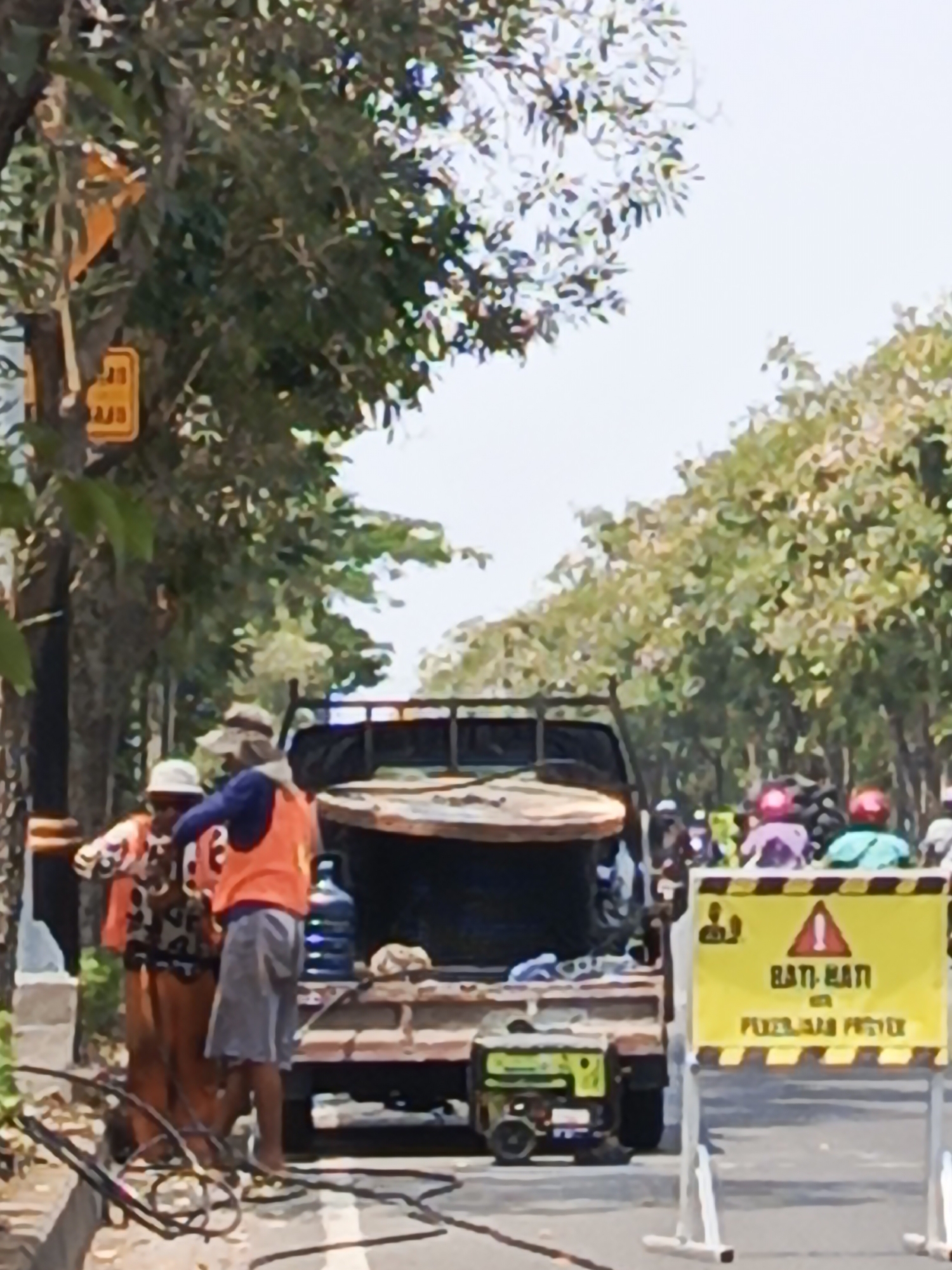 A low-angle, slightly blurred medium shot captures two individuals working on a roadside in Surabaya, Indonesia. The scene appears to be a construction or repair site, indicated by a yellow and black striped barrier sign in the foreground that reads "HATI-HATI DI PERKERASAN PROYEK" (Caution on Construction Pavement).
To the left, a person wearing a white bucket hat and a patterned shirt is interacting with another individual who is wearing a blue long-sleeved shirt and an orange vest, and grey shorts. They appear to be engaged in some sort of task, possibly related to the electrical cables and equipment visible on the ground. A large blue water jug is present on the back of a flatbed truck, positioned behind the individuals.
The truck itself is loaded with what appears to be a large, flat, rounded object, possibly a section of concrete or a similar construction material. A green generator is also visible on the truck bed. In the background, several motorcycles with riders wearing brightly colored helmets are lined up, suggesting traffic or the presence of other workers. The environment is characterized by a tree-lined street with dappled sunlight filtering through the leaves. The sky is bright and overcast.