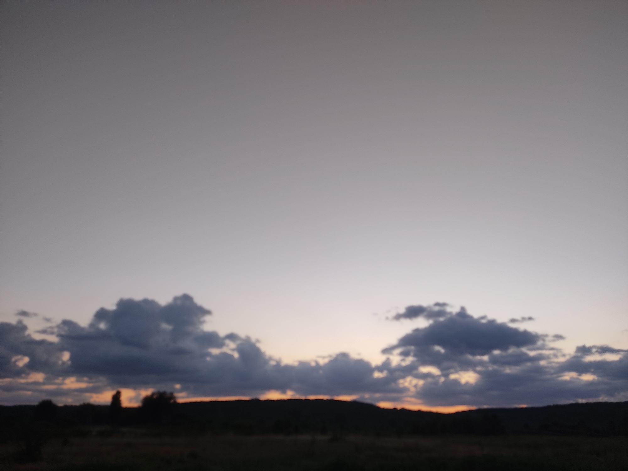 This is a photograph of a landscape at dusk in Cherevky, Ukraine. The sky is a soft gradient of pale gray at the top, transitioning to a light pink and orange hue near the horizon. Dark, textured clouds are scattered across the middle ground, some appearing heavy and shadowed, others catching the fading light. Below the sky, a silhouetted, hilly terrain is visible, with dark shapes of trees and vegetation dotting the landscape. In the far distance, the faint outline of a man-made structure or a prominent tree can be discerned against the horizon. The foreground consists of a darker, blurred area that suggests fields or uneven ground. The overall mood is tranquil and serene, characteristic of twilight.